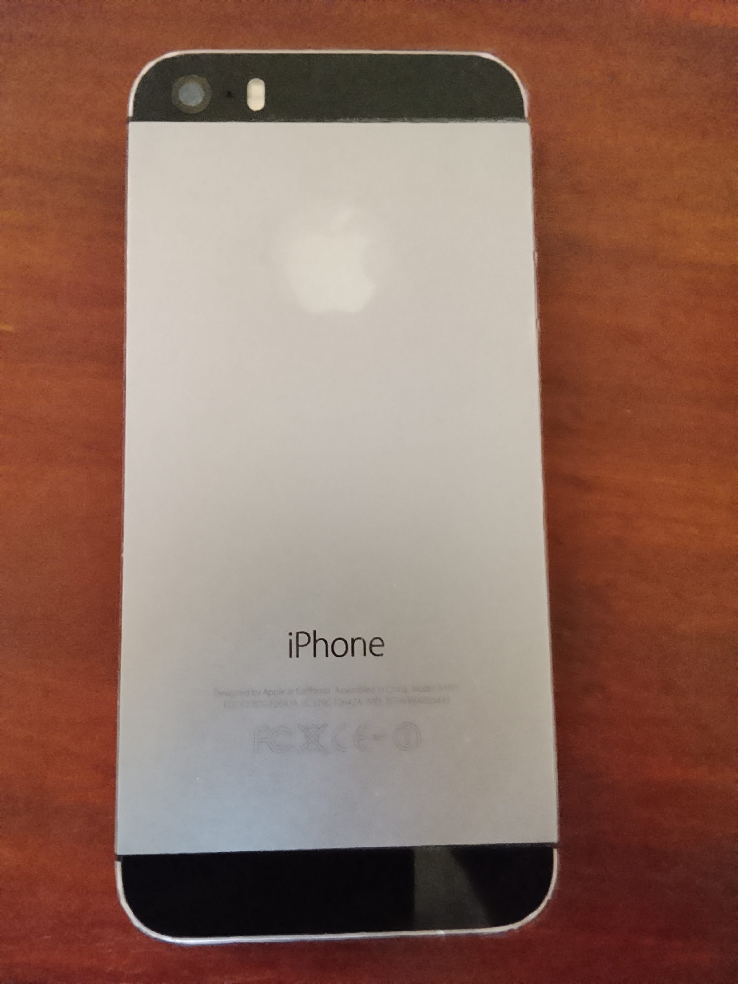 A silver iPhone 5 is photographed from behind, lying on a wooden surface. The phone is oriented vertically with the camera at the top. The Apple logo is visible in the center of the phone's back. Text indicating "iPhone" is present below the logo, followed by regulatory information. The background consists of a reddish-brown wooden table with a visible grain. The image does not provide any information about Karangklesem, Indonesia.
