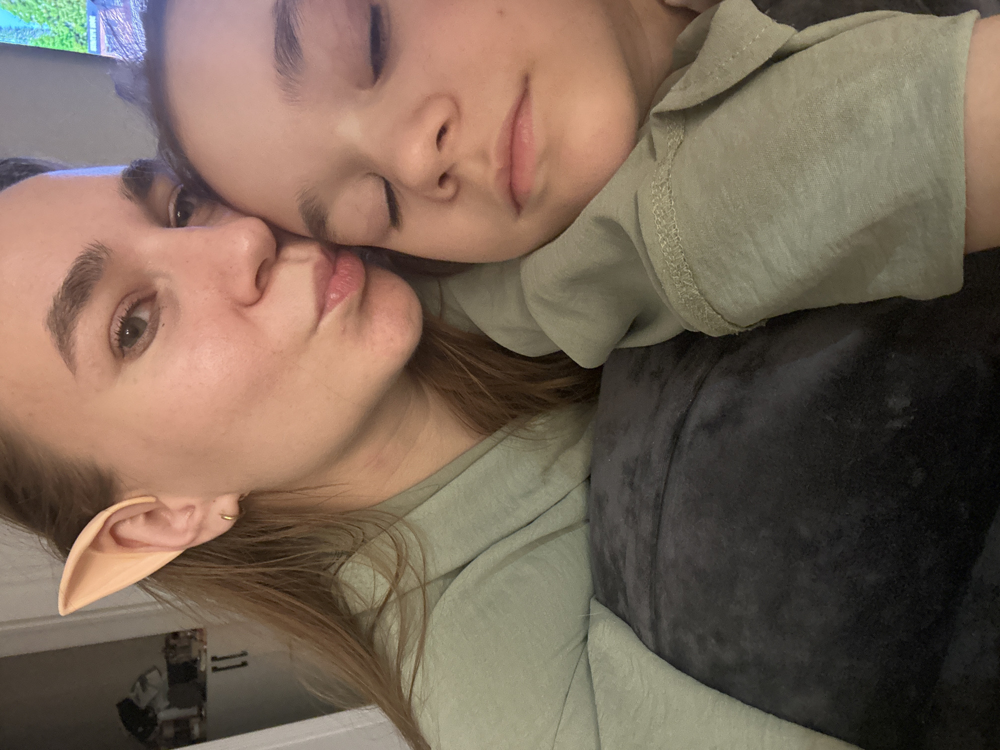 Two people are visible in the image. One is awake with one brown eye fully visible, wearing a prosthetic elf ear and a light green top. The other is asleep with their eyes closed, wearing a similar light green top. They are lying close together. The background is a plain gray wall, and there is a glimpse of a room with a wardrobe in the lower left corner. A TV is visible at the top of the image. There is a dark gray velvety fabric draped around them. The image is rotated sideways. The location is not discernable from the visual information provided.