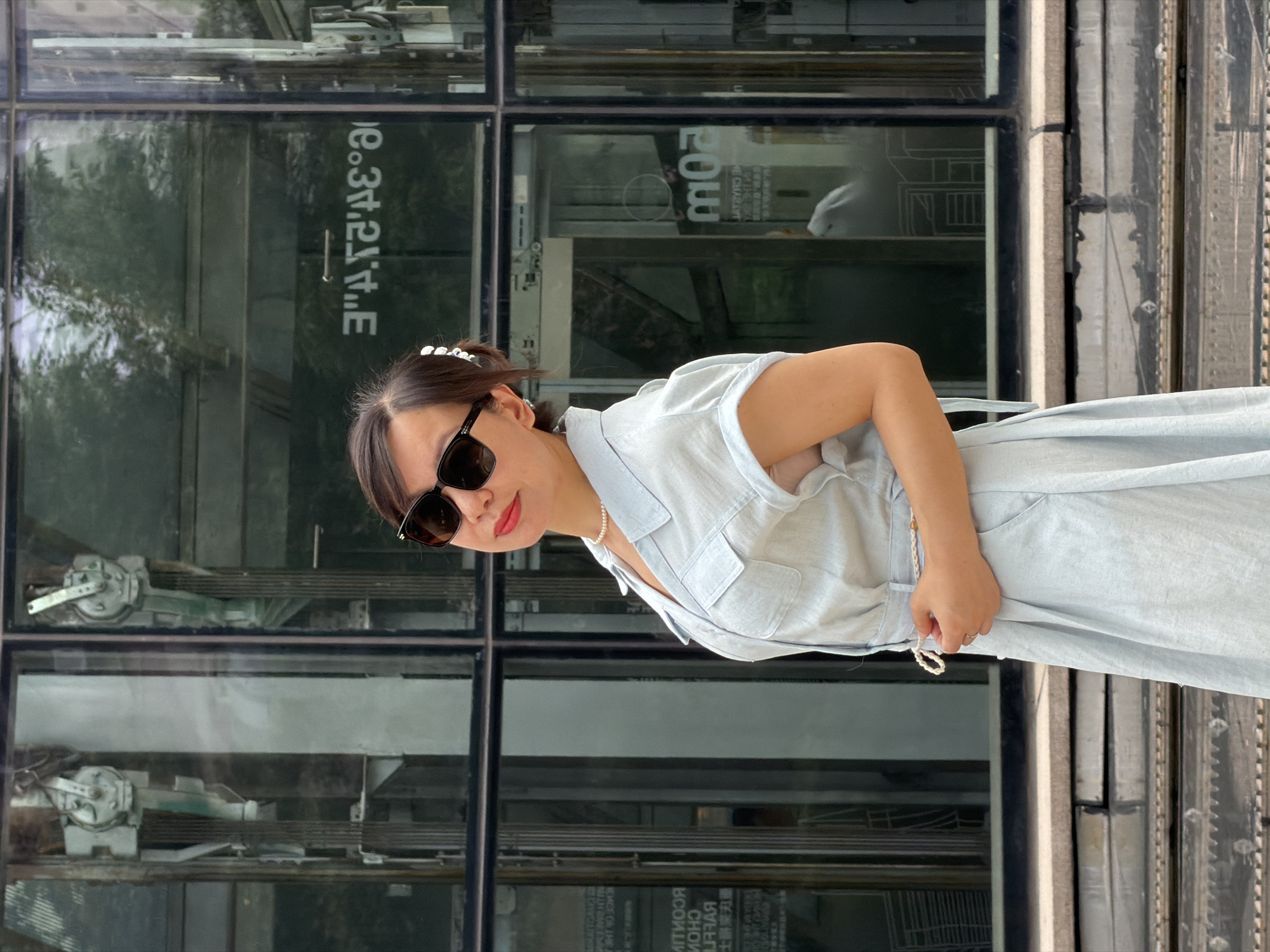 A woman with dark hair, wearing sunglasses, a light blue collared dress, and a pearl necklace, stands in front of a modern building with large glass windows. The building appears to be in Yuzhong District, China, as evidenced by reflections on the glass showing signage that includes Chinese characters and what seems to be a building directory or map. The woman is positioned slightly to the left of the frame, looking towards the camera with a subtle smile. Her right hand is near her waist, touching the braided belt of her dress. The background reveals the architectural details of the building, including elevators and metal structures within the glass facade, as well as some greenery reflected from outside. The ground in front of the building features a textured surface, possibly concrete or stone steps, with metallic elements underneath.