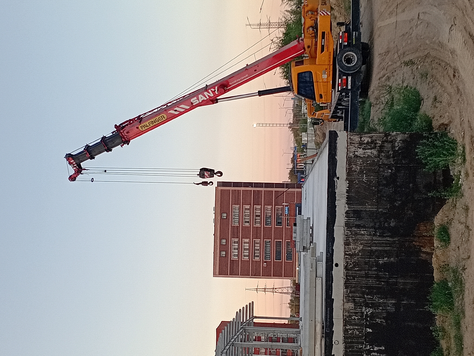 In Semey, Kazakhstan, at a construction site under a clear, light blue sky transitioning to a warm orange at the horizon, a large yellow SANY mobile crane is positioned to the right of the frame. The crane's extended red boom reaches upwards and to the left, with the brand "PALFINGER" and "SANY" visible on its side. Two large hooks hang from the crane's upper section. In the mid-ground, a multi-story brick building with rows of windows stands to the left of center. To the left of this building, a partially constructed metallic structure with a framework is visible. In the foreground, a flat concrete surface is covered with several long, stacked concrete slabs. The ground around the construction site consists of dirt and patches of green vegetation. The scene depicts ongoing construction work in an urban environment.