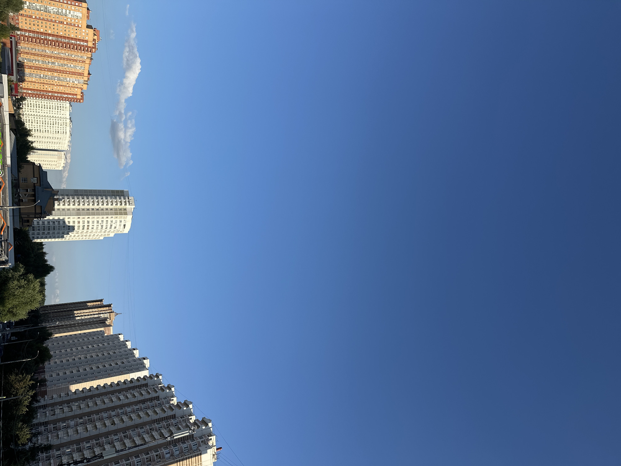 The image captures a street-level view of residential buildings in Podolsk, Russia, under a clear blue sky. Several multi-story apartment buildings dominate the scene, with one particularly tall, white building standing prominently in the center. To the left, two orange-toned buildings are visible, featuring numerous balconies and windows. In the distance, more buildings line the horizon, some angled towards the viewer. Green trees and vegetation dot the lower portion of the image, interspersed with streetlights and power lines, suggesting an urban environment. A few wispy clouds are present in the sky. There are no people or visible actions in the frame; the scene appears static and devoid of human activity. The lighting suggests it is daytime, likely late afternoon given the angle of the shadows.