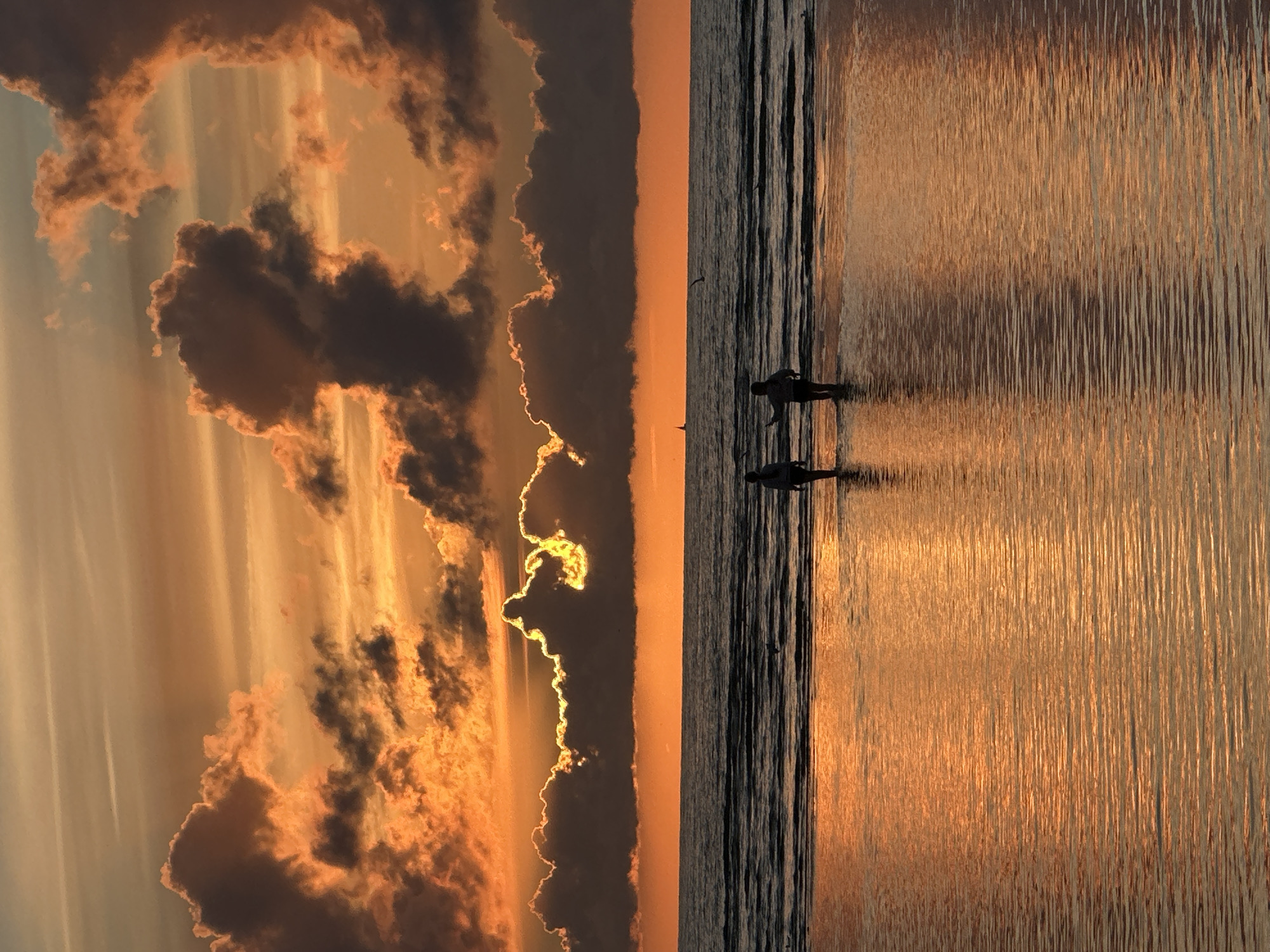 The image depicts a serene sunset scene on a beach. Two figures, silhouetted and appearing to be children, are walking in shallow water near the shore. The sky is dramatically lit by the setting sun, with vibrant orange and yellow hues painting the clouds. The ocean is calm, with gentle waves reflecting the colorful sky. The foreground shows the wet sand reflecting the sunset, creating a shimmering effect. The image is a vertical composition, with the sky occupying the upper half and the ocean and beach occupying the lower half. The scene does not appear to be located in Tallinn, Estonia, as there are no identifiable landmarks or architectural features to suggest this location.