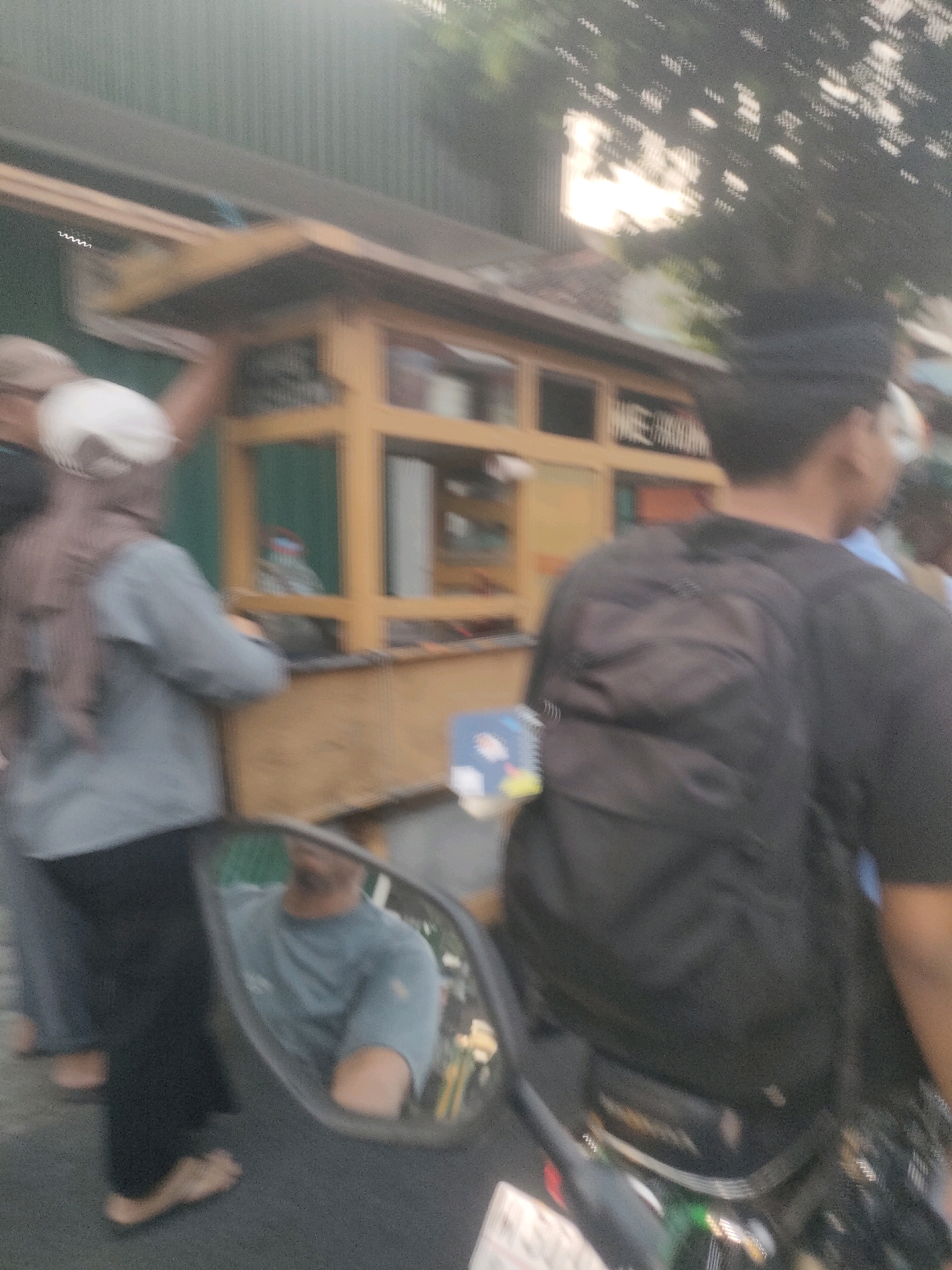 A blurred image depicts a street scene in Surabaya, Indonesia, with a focus on a food stall and several people. A woman wearing a headscarf and grey top stands at the stall, which is constructed of light-colored wood and has shelves displaying items. To her right, a person wearing a black backpack and dark clothing is partially visible, facing away from the camera. In the foreground, a motorcycle side mirror reflects the image of a person wearing a grey t-shirt. The background shows a building with green siding and dappled sunlight filtering through trees. The overall impression is of a busy urban environment.