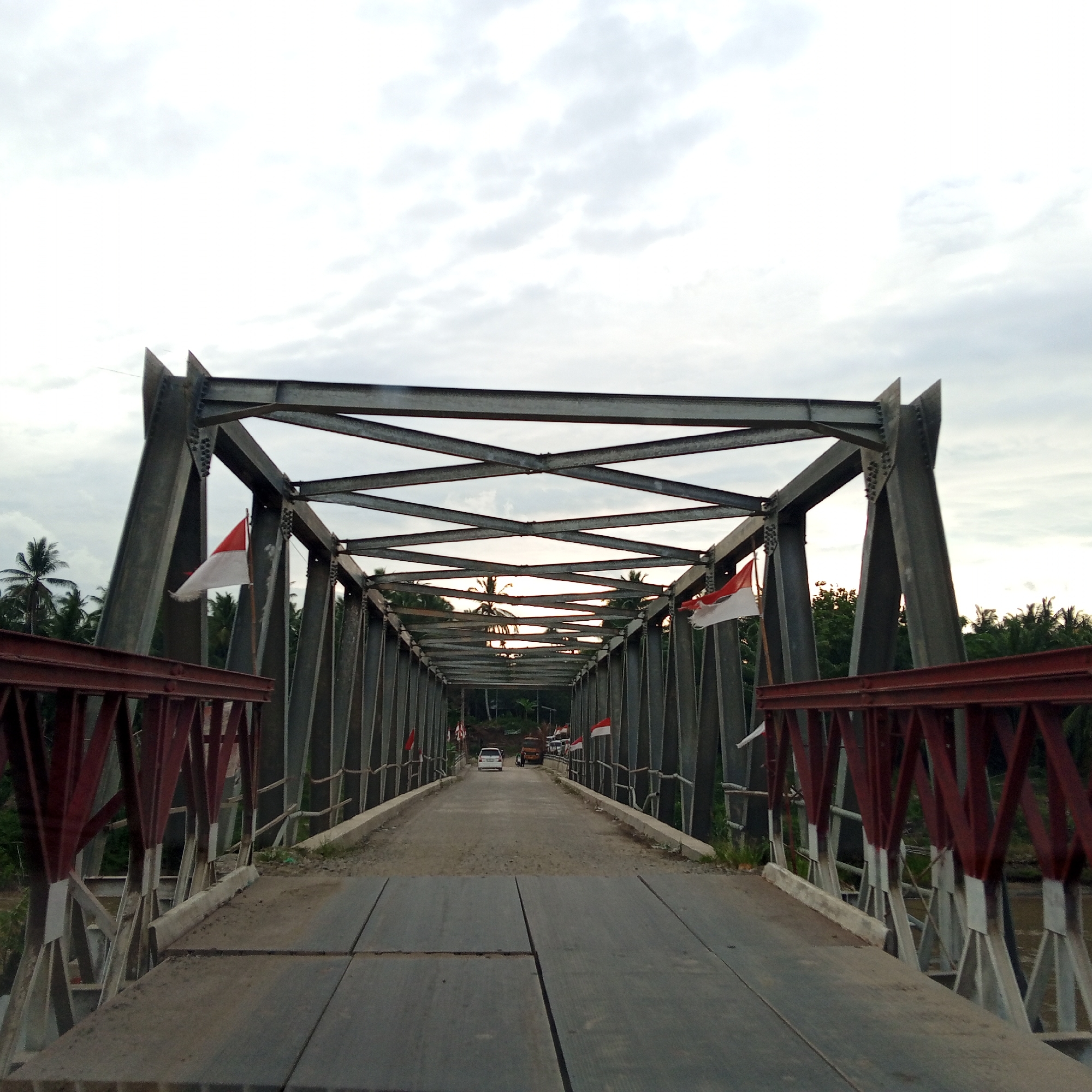 This image captures a frontal, slightly elevated perspective of a metal truss bridge in Teupinreudeup, Indonesia, extending into the distance under an overcast sky. The bridge's primary structure consists of dark grey metal beams, forming an overhead lattice and vertical-diagonal trusses along the sides, connected by rivets or bolts. Prominent reddish-brown metal railings and supports are visible on the immediate left and right in the foreground, bordering the bridge entrance. The bridge deck starts with flat, rectangular grey panels, transitioning further along its length into a dirt and gravel road surface. Multiple red and white flags, emblematic of Indonesia, are affixed to the internal bridge trusses on both sides, some exhibiting signs of wear. Several vehicles are present on the bridge road, including a white car, a light-colored truck, and other less distinct vehicles further away, all oriented away from the camera. Dense tropical vegetation, featuring numerous palm trees and other broadleaf trees, flanks both sides of the bridge, and a murky brown body of water is partially visible beneath the bridge structure on the right.