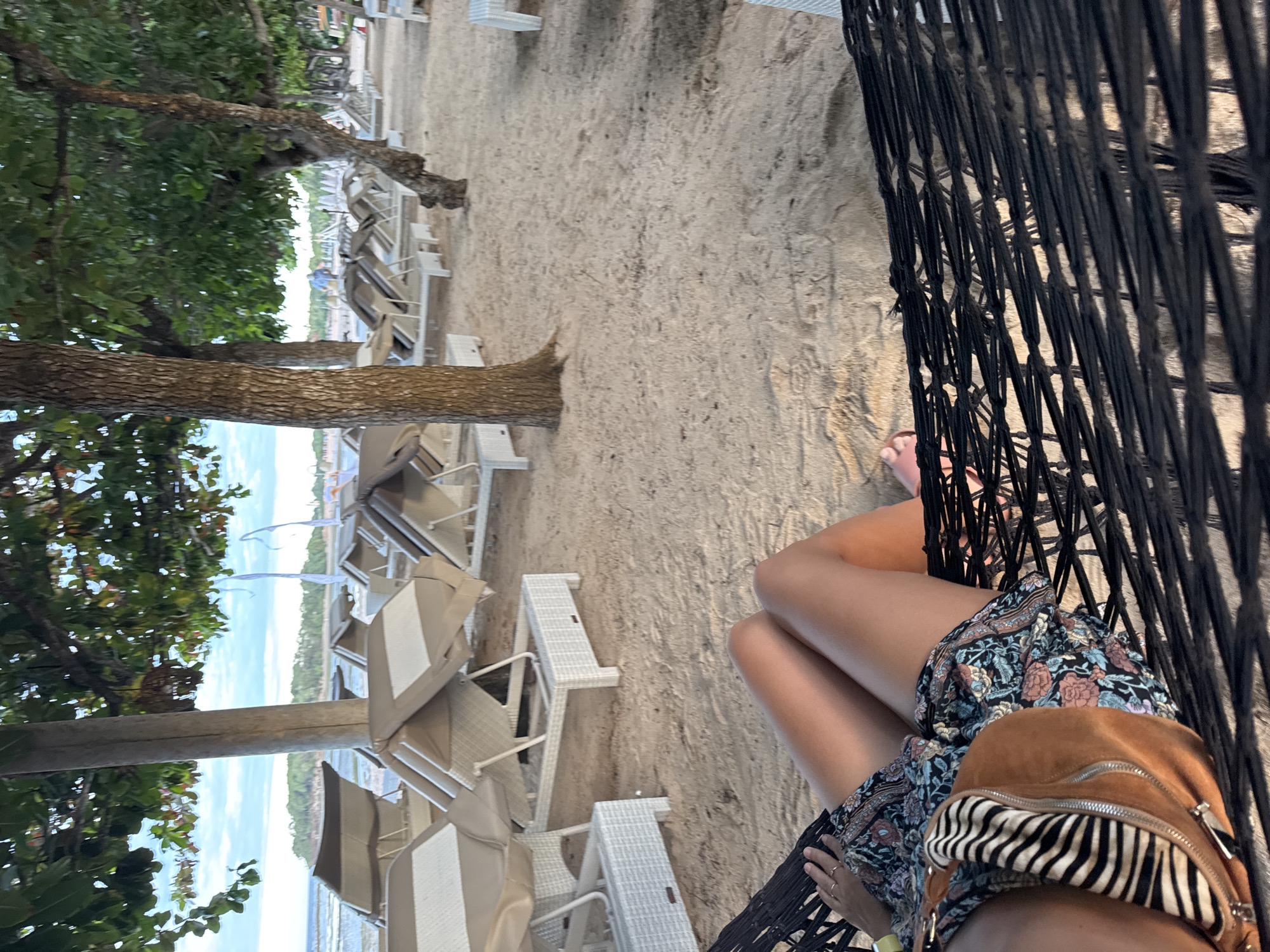 A person is sitting in a black woven hammock on a sandy beach in Unknown, Unknown. The person is wearing a floral dress and has a brown bag with a zebra-striped accent. Their legs are visible, and they are holding onto the hammock. In the background, there are beach chairs lined up on the sand, and some trees are visible on the left side. The overall scene depicts a relaxed and scenic beach setting.
