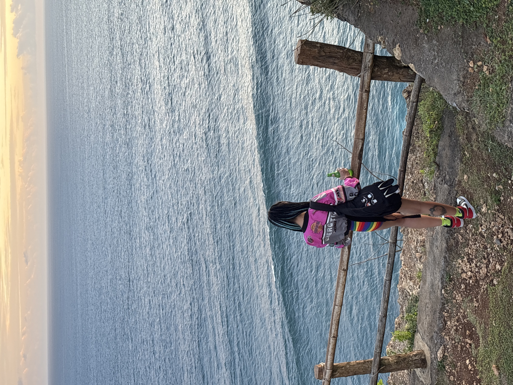 A long shot shows a person perched on the edge of a cliff overlooking a vast ocean, possibly at Uluwatu Temple, a scenic cliffside Hindu temple in Kuta Selatan, Indonesia. The person stands behind a crude, wooden fence, holding a green bottle. The person is wearing a pink jacket, black shorts, and rainbow-striped socks, with red and white sneakers. They also have a black backpack over their shoulder and blue streaks in their black hair. The cliff edge is rocky with patches of grass. The ocean is calm, with a light sunset glow on the horizon.