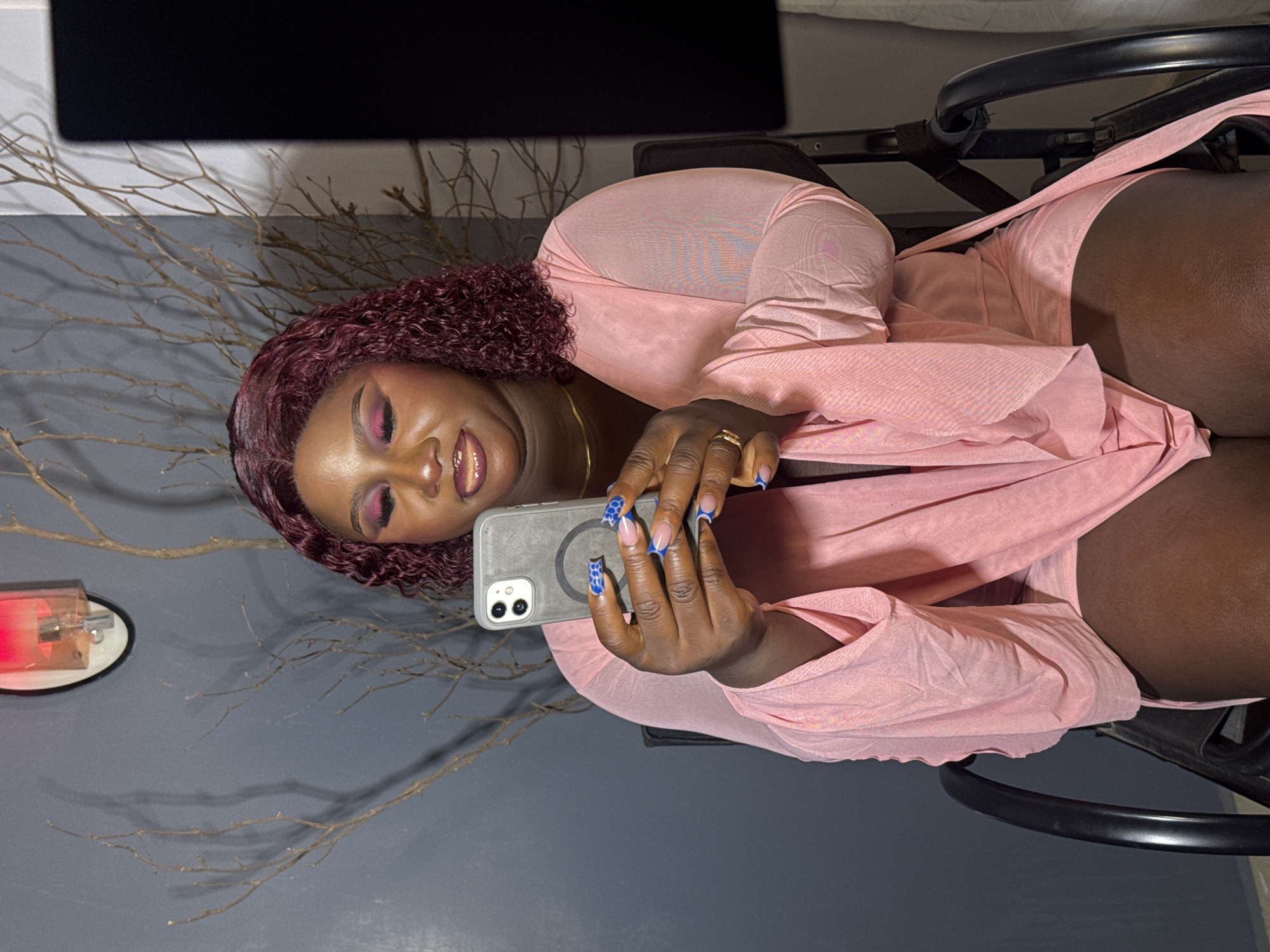 The media file shows a close-up, portrait-style image of a dark-skinned woman with curly, dark red hair and striking pink eye makeup. She is looking at her phone, which she is holding in her hands, and appears to be taking a selfie or recording a video. The woman is wearing a light pink, sheer, flowing garment that drapes over her shoulders and body. A gold necklace is visible around her neck.
The setting appears to be indoors, with a dark gray wall in the background. To the left of the woman, a decorative element resembling dry branches or twigs is visible. There is also a small, cylindrical object with a red light on it. The overall mood of the image is relaxed and intimate, with the woman seemingly engrossed in her phone. The lighting is soft, suggesting it might be evening or a dimly lit indoor space. No text is visible in the image. The woman's hands are adorned with long, blue-tipped nails, adding a pop of color and detail. She is wearing a ring on her right hand. The image is shot from a slightly elevated angle, looking down at the woman.