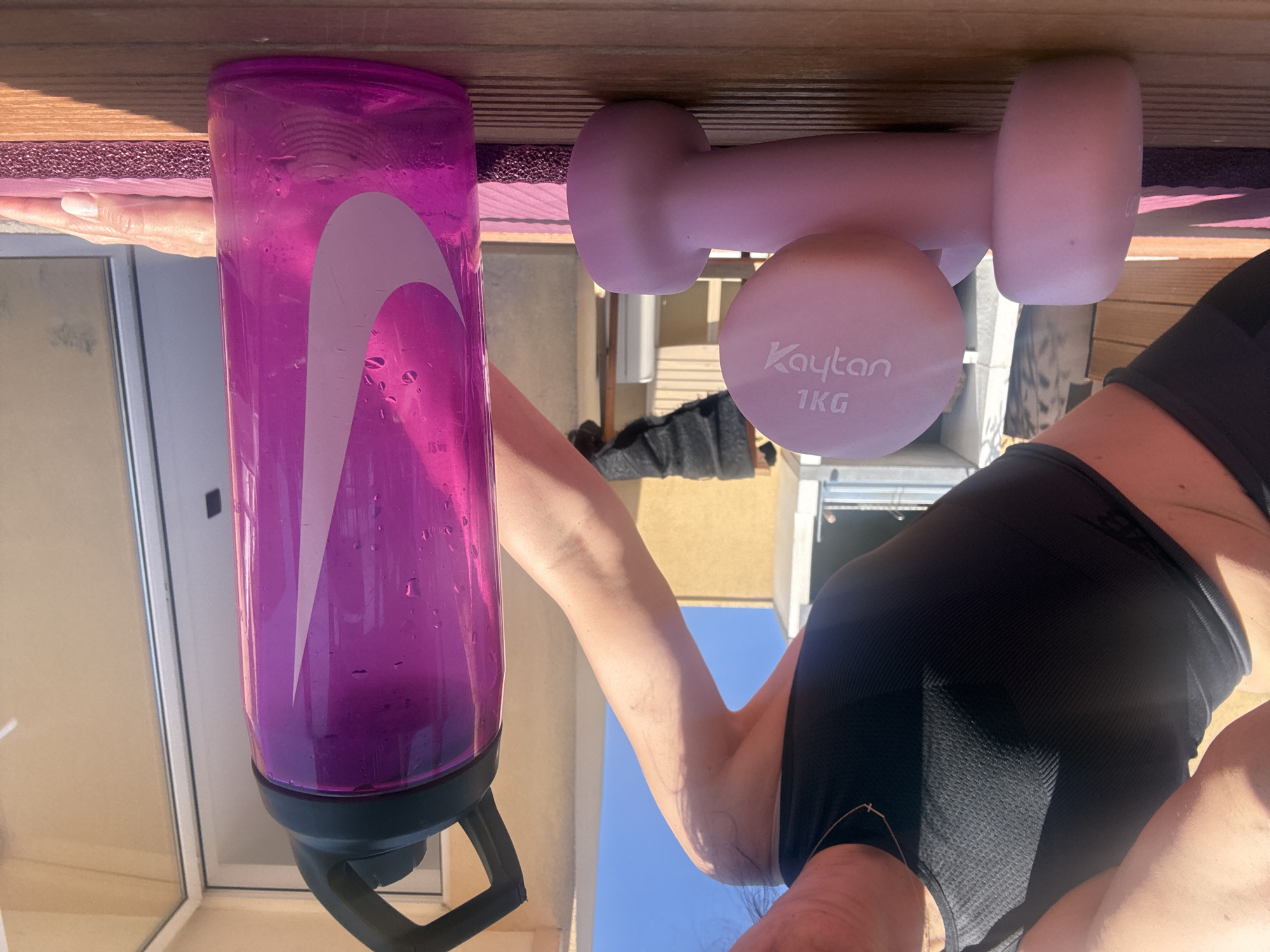 A purple Nike water bottle and a pink 1 kg Kaytan dumbbell sit on a maroon yoga mat on a wooden surface. A person with tan skin and wearing a black tank top and shorts is visible. The background shows parts of a building, including a window and exterior elements, against a blue sky. The location is Vila Nova de Gaia, Portugal.
