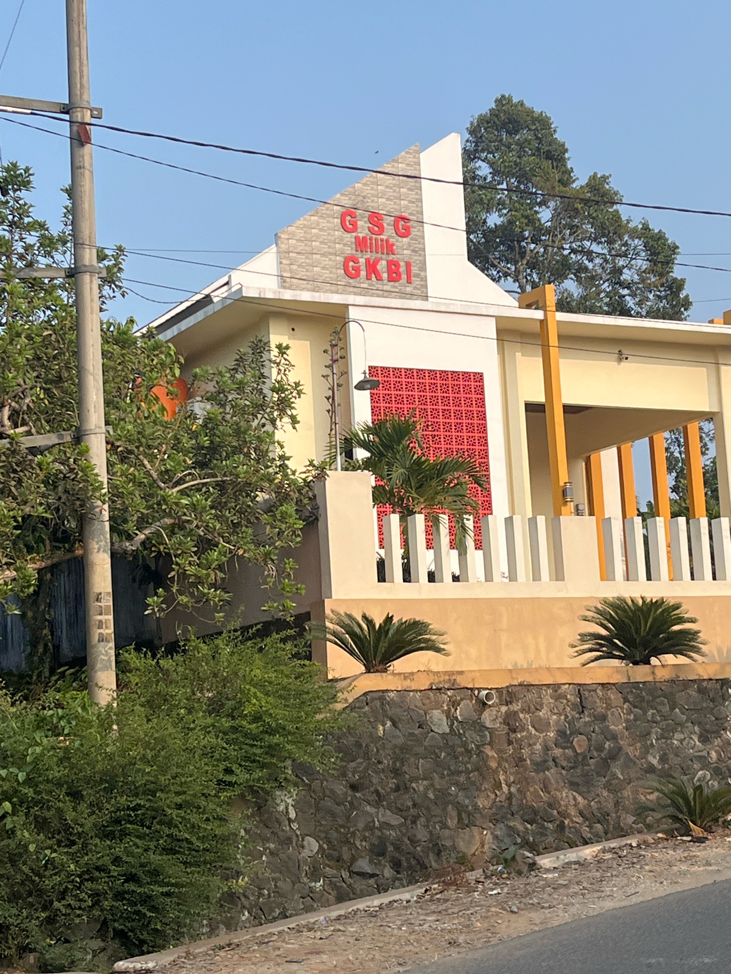 The image shows a building with the red lettering "GSG MILIK GKBI" on its facade. The building is painted in yellow and white, with a decorative red patterned panel on one side. There is a fence in front of the building and some palm trees. A street is visible in the foreground. The building appears to be located in a residential area with vegetation. The context provided states that the location is the city of Margodadi, Indonesia. No people are visible in the image.