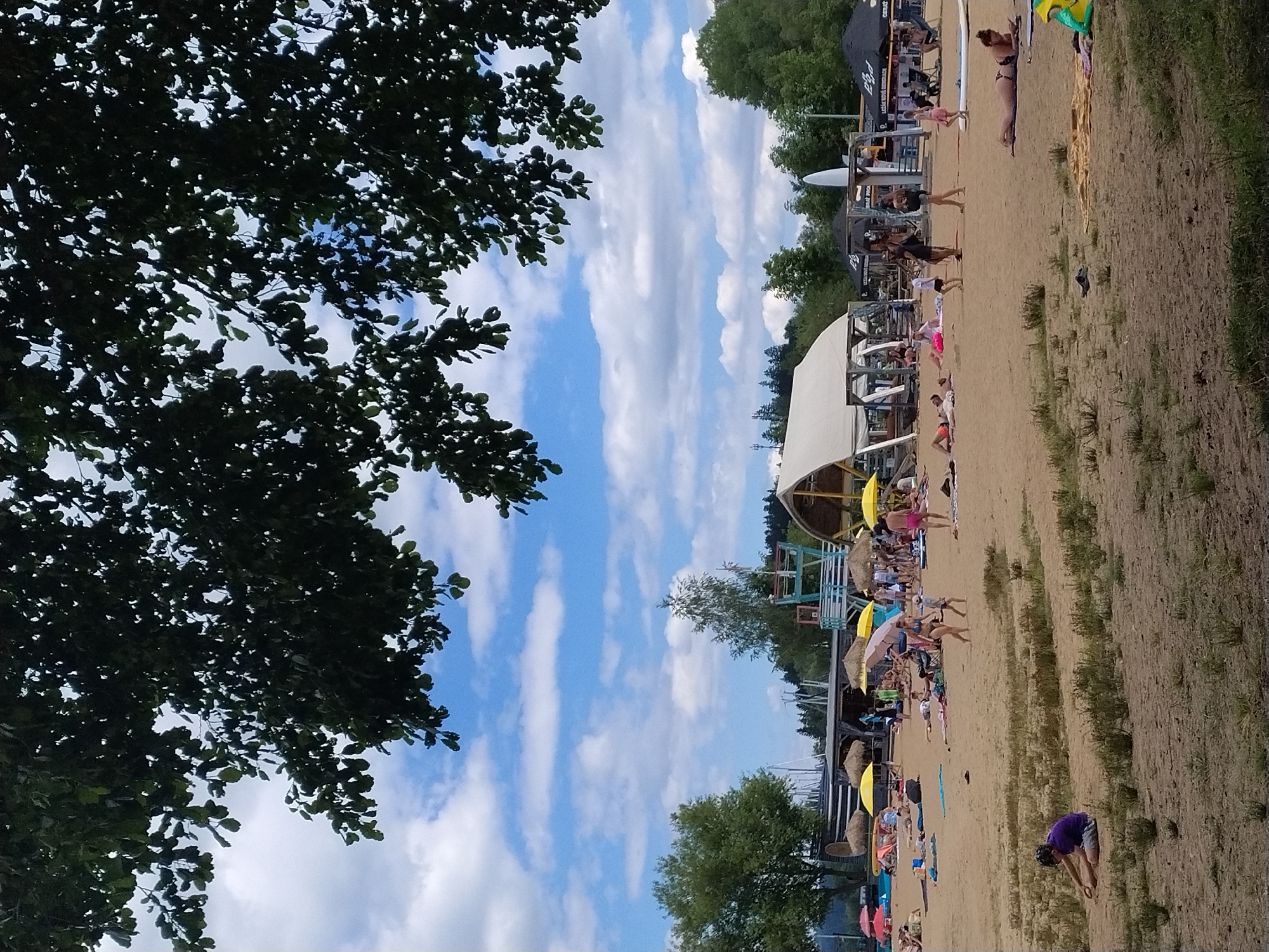 The image captures a sunny day at a beach resort in Radslav, Czechia. The scene is filled with people enjoying various activities. In the foreground, a person in a purple shirt is sitting on the sand. Further back, a diverse group of individuals are scattered across the beach, some sunbathing, others walking, and children are playing. Beach umbrellas, both yellow and straw-colored, provide shade. There are several structures on the beach, including a large white tent-like building and a lifeguard stand. In the background, lush green trees line the horizon, and a clear blue sky with scattered white clouds serves as the backdrop. The presence of beach chairs, towels, and what appear to be food or drink vendors suggests a well-equipped recreational area.
