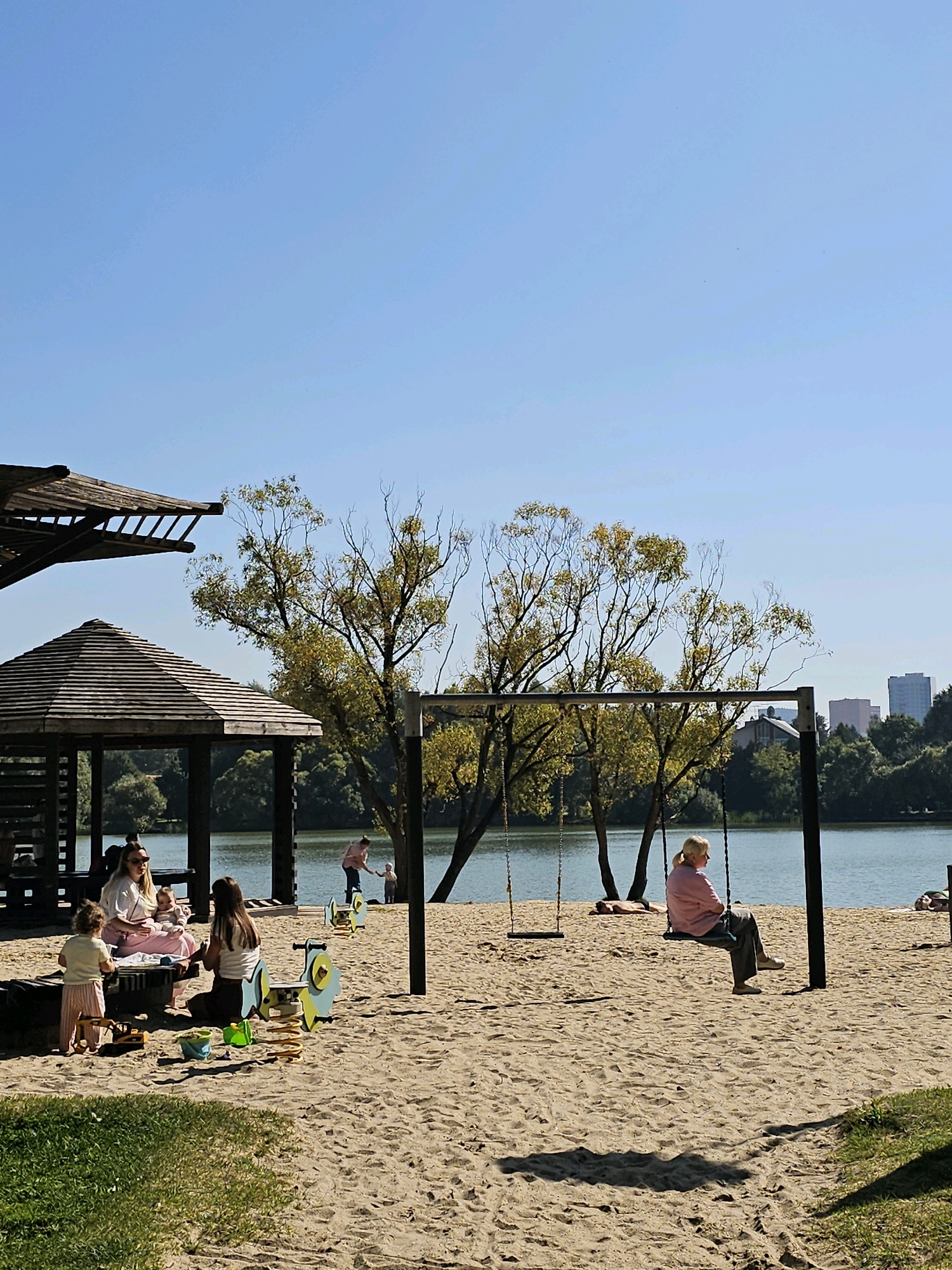 The image depicts a sandy playground area situated beside a body of water, likely a lake or river, under a clear blue sky. In the foreground, children and adults are present. A woman in a light-colored top and pink skirt sits with a baby on her lap at a picnic table, while a small child in striped pants stands nearby. To their right, another woman sits facing away from the camera. In the mid-ground, a swing set with two swings is visible, with an older woman on one of the swings. Further in the background, trees line the water's edge, and beyond that, a cityscape with buildings can be seen. The scene suggests a recreational area, possibly a park or beach, in Moscow, Russia.