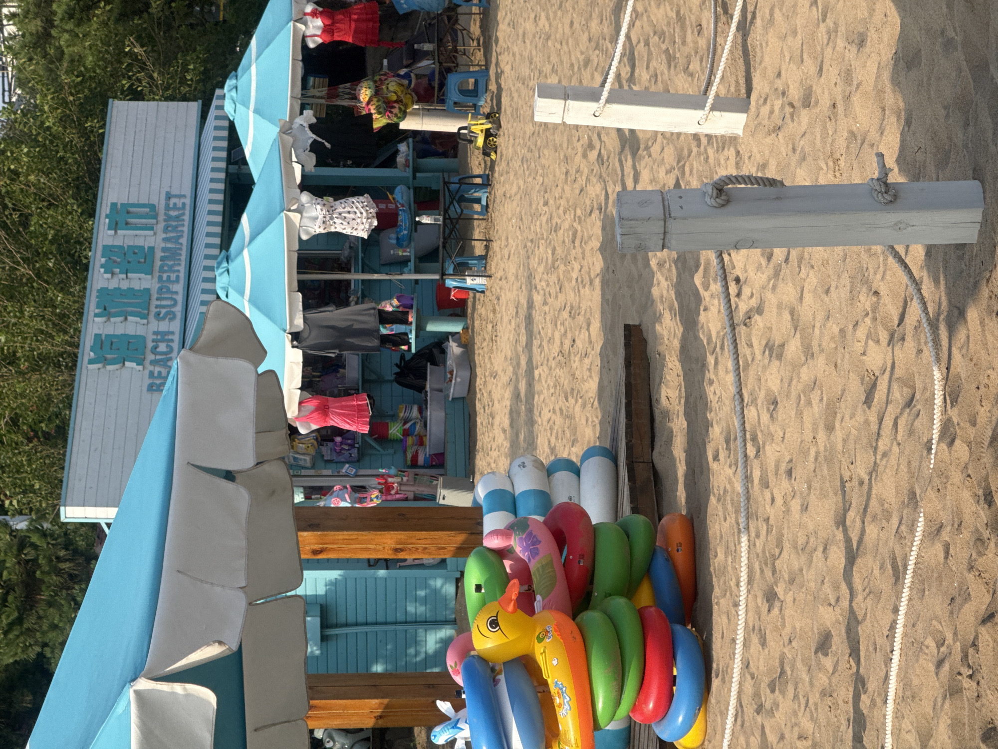 This image depicts a scene on a sandy beach, likely in Qinhuangdao, China, given the presence of Chinese characters on signage. In the background, a storefront with a light-colored facade and blue lettering reads "BEACH SUPERMARKET" in English, with corresponding Chinese characters above. The store appears to be a kiosk or small building, with blue awnings and umbrellas providing shade. Merchandise, including clothing like dresses and shirts, is displayed.
In the foreground, a colorful stack of inflatable swimming rings, including a yellow duck-shaped ring, is positioned on a wooden platform. Behind the rings, more inflatable objects and stacked items are visible, along with a small yellow toy construction vehicle on the sand. Two wooden posts with ropes stretched between them mark a boundary on the beach. The sand is rippled and shows shadows, indicating a sunny day. No people are clearly visible in the image.