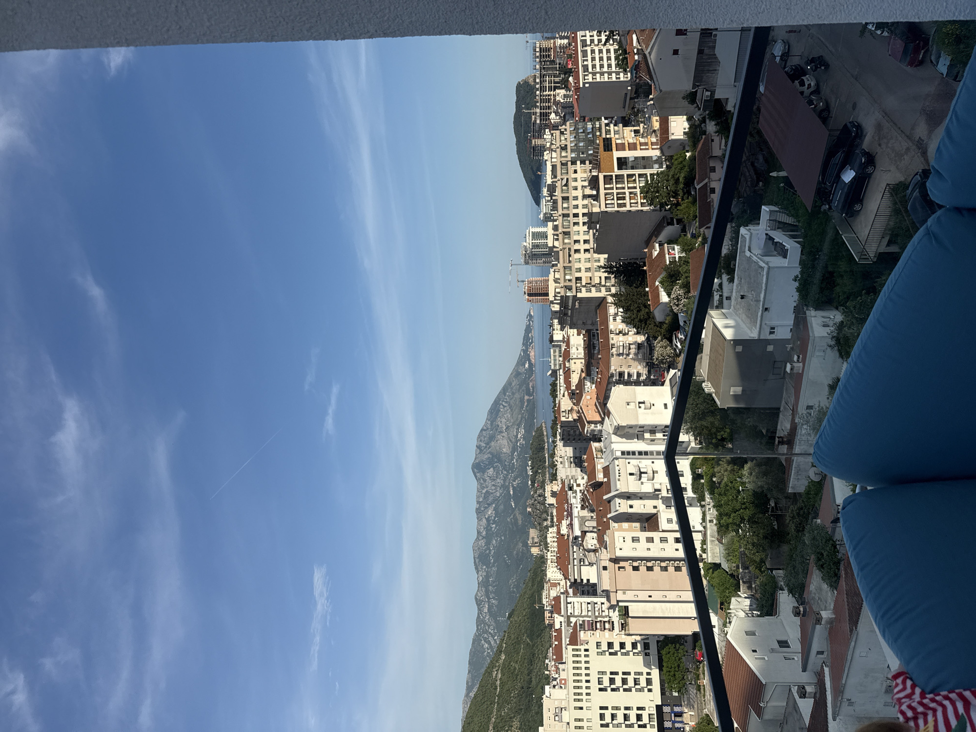 The scene is a high-angle, outdoor view in Budva, Montenegro. The city is visible, composed of light-colored buildings with varying roof colors. There's a mountain range in the background, separating the city from the blue sky with some clouds. The Adriatic Sea is situated between the mountains and the city. The image is taken from a high vantage point, possibly a balcony, as a blue cushion and a railing are visible in the foreground. The city appears densely built with a mix of residential and commercial structures. Roads and parked vehicles are also visible.