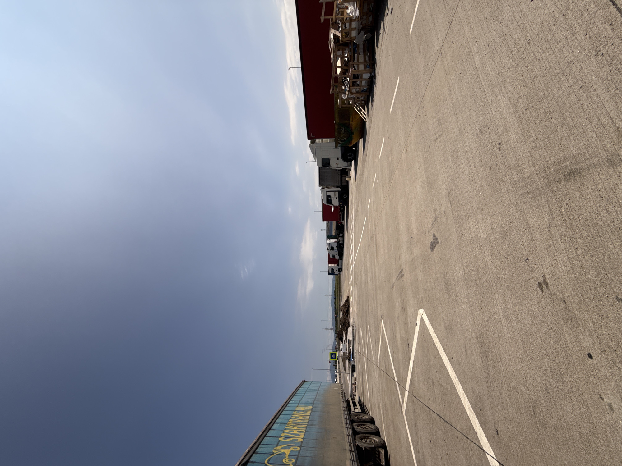 An outdoor, low-angle, full-shot image captures a truck parking lot under a clear blue sky with a few wispy clouds in the distance. The foreground is dominated by a concrete surface with white directional and parking lines. A large blue truck with yellow lettering that reads "SZAKITRANS.HU" is parked on the left side of the frame, its wheels and tires visible. In the mid-ground, a line of semi-trailer trucks, predominantly white and red, are parked. Some of these trucks have visible company logos or text. A yellow dumpster and piles of wooden debris are also present in the mid-ground. In the far distance, utility poles and a treeline can be seen, with mountains forming a backdrop. The scene appears to be a rest stop or a depot for commercial vehicles, located in the vicinity of Sanpetru, Romania. No people are visible in the frame. The lighting suggests it is daytime, with the sun high in the sky.