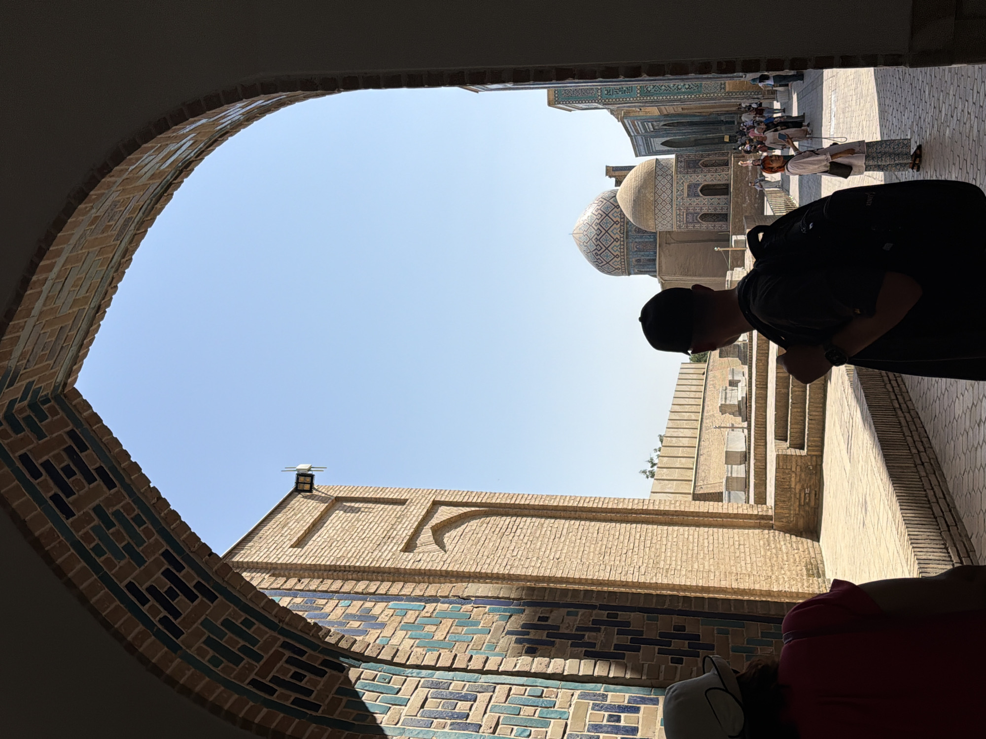 This image captures a view through an arched entryway at the Registan complex in Samarkand, Uzbekistan. The entryway is decorated with intricate geometric patterns of brown, blue, and teal tiles. To the left, a brick wall with similar tiled accents is visible. In the background, under a clear blue sky, several domed structures characteristic of Registan's madrasahs are prominent. The foreground features the backs of two individuals, one wearing a dark baseball cap and backpack, and another with a white hat and red shirt. Further in the background, a group of tourists are visible, some taking photographs. The ground is paved with cobblestones.
