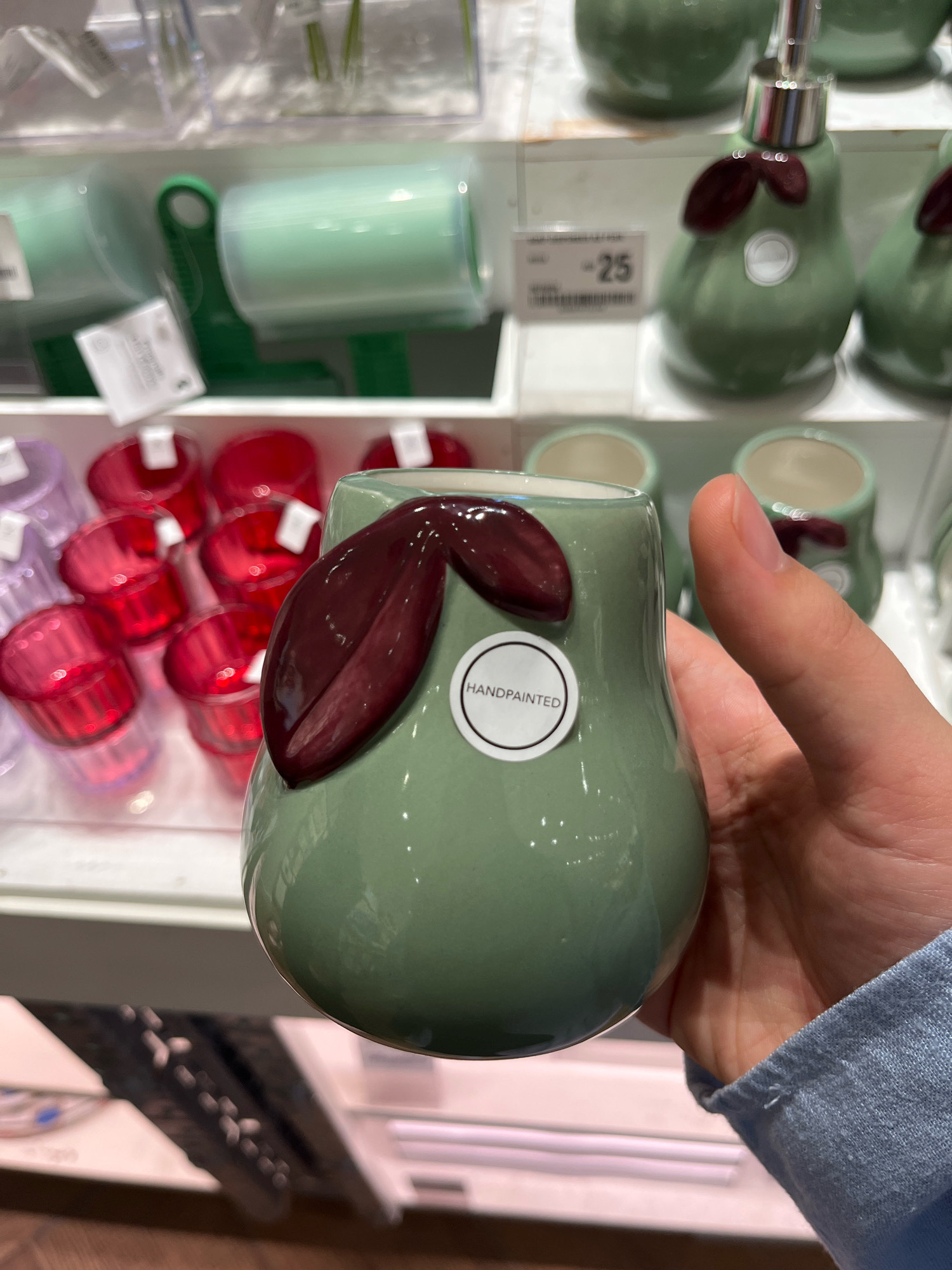 The image shows a close-up of a hand holding a light green, pear-shaped ceramic object, possibly a small vase or dispenser, adorned with two dark red leaf-like embellishments. A circular white sticker on the object reads "HANDPAINTED". In the background, shelves are visible stocked with various items, including red glass containers, a green lint roller, and other pear-shaped ceramic items. The setting appears to be a retail store. The background also shows a price tag with the number "25". The image quality is slightly blurred, suggesting it was taken in a retail environment, potentially in Dubai, United Arab Emirates, given the context provided.
