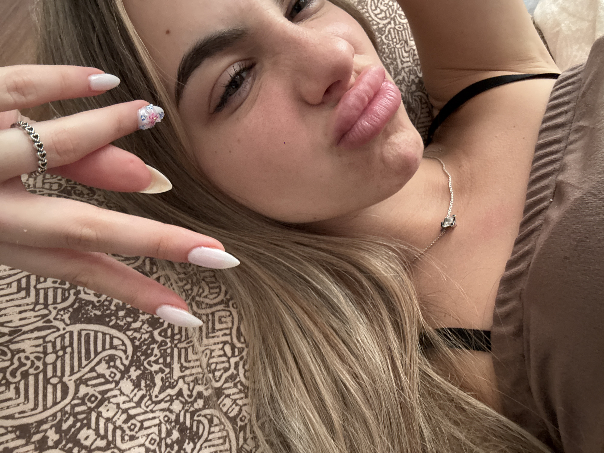 A close-up, eye-level shot features a young woman lying down, looking directly at the camera with a playful, pouting expression. Her head is tilted, and her long, straight blonde and light brown hair cascades around her. Her face is partially visible, focusing on her eyes, nose, and full, pink lips which are pursed. She has defined eyebrows and visible eyelashes.
Her left hand is raised towards her face, showcasing long, pointed, white acrylic nails. One nail, on her ring finger, features a small, intricate floral design in pink and blue. A silver ring with a pattern of connected hearts adorns her ring finger. Around her neck, a delicate silver chain necklace with a small, square, possibly jeweled pendant is visible.
She is wearing a thin black strap, likely from a bra or camisole, and a light brown or taupe top with some ruffled or gathered texture. The background consists of a patterned pillow or blanket with an ornate, possibly geometric or tribal design in varying shades of brown and beige. The setting appears to be indoors, suggesting a relaxed environment like a bedroom. There is no visible text.