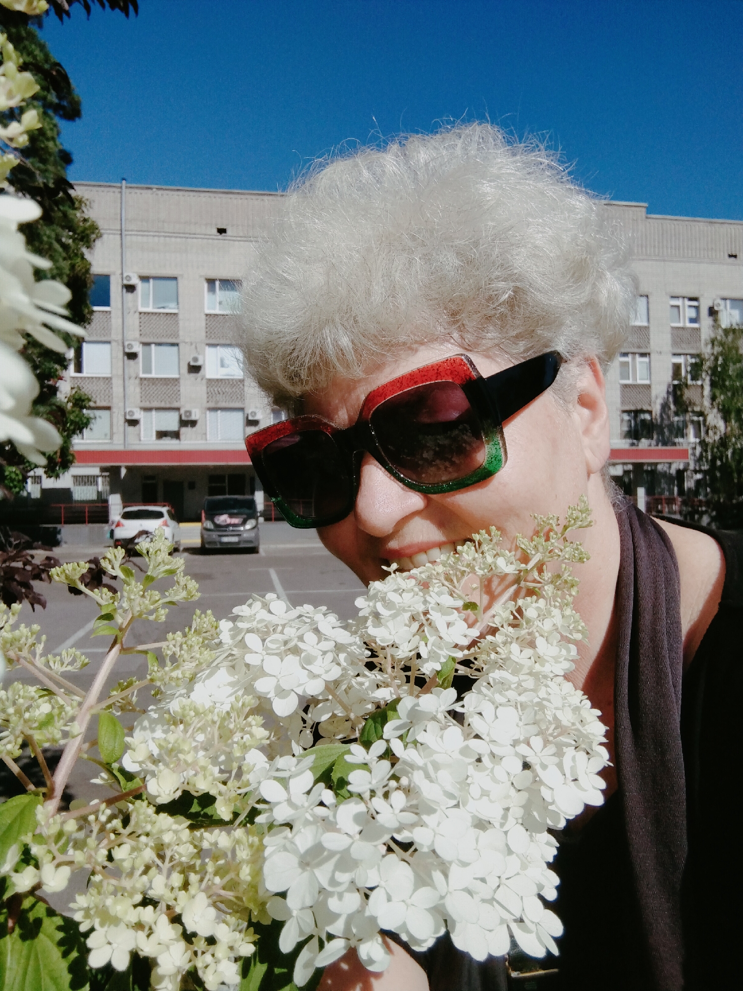 A woman with white, permed hair and large sunglasses with red and green frames is holding a bouquet of white flowers in the foreground. She is wearing a dark top and is partially obscured by the flowers, with only her face and shoulder visible. Behind her, a light-colored, multi-story building with numerous windows and air conditioning units is visible. The building appears to be in a city setting, with cars parked in front of it. Trees are also present in the background. The sky is clear and blue. The image is taken outdoors on a sunny day, and it is likely located in the city of Horishni Plavni, Ukraine, given the context.