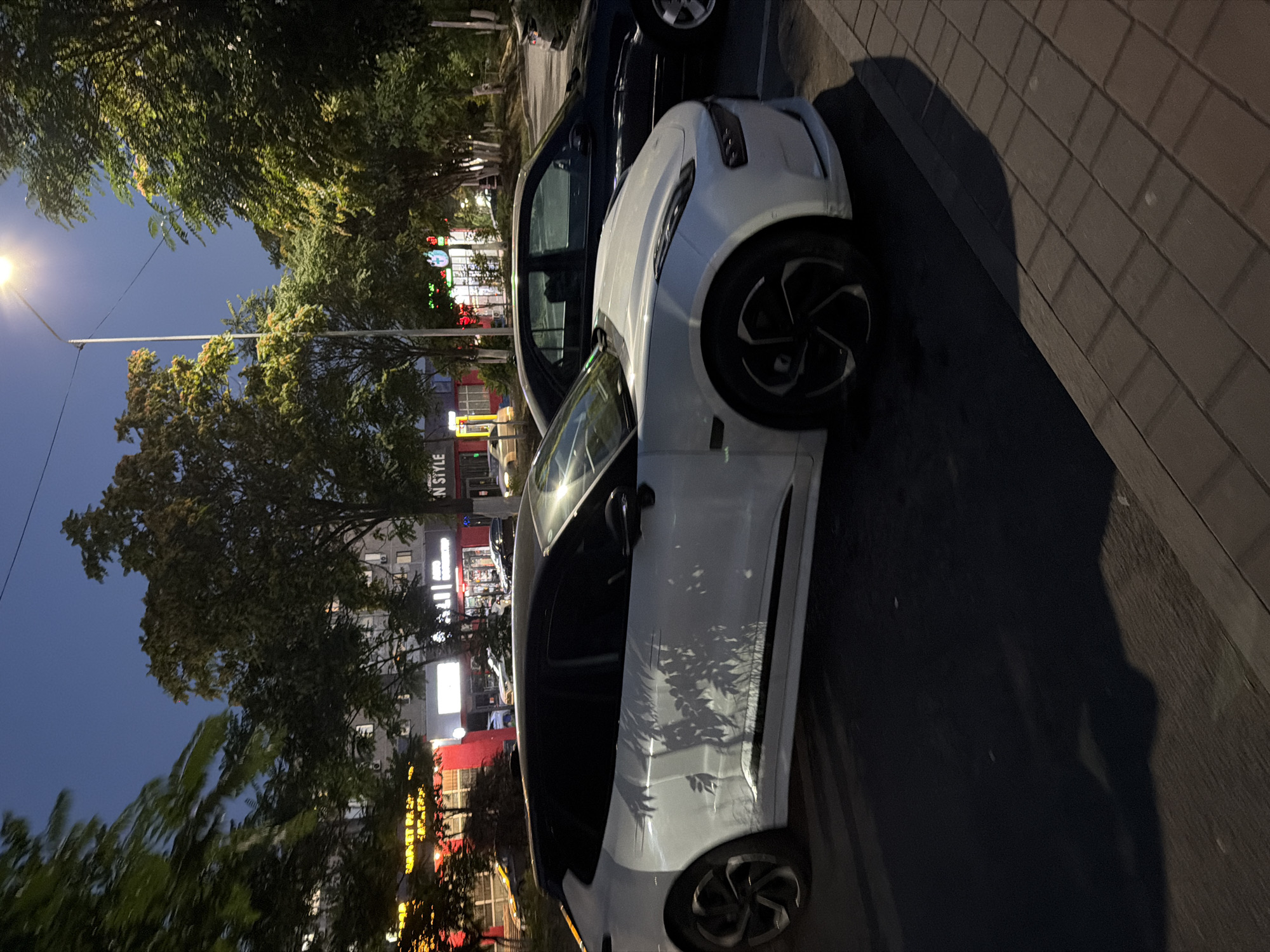 The image shows a white car parked on a paved sidewalk at dusk. The car is the primary subject, and its side profile is visible. It has a modern design with sleek lines and a distinctive wheel. A dark-colored car is parked behind it, mostly out of focus.
The scene is set on a street lined with trees, casting shadows on the car and the sidewalk. In the background, buildings with illuminated signs are visible, suggesting an urban or commercial area. One sign, partially obscured by trees, appears to have text in a foreign language, possibly Cyrillic. Another sign, clearer in the crop, reads "N STYLE". The overall lighting suggests it is evening, with artificial lights from the buildings and streetlamps starting to illuminate the area.
The weather appears calm, with no indications of rain or wind, though some leaves on the trees are blurred, possibly due to a slight breeze or camera movement. The mood is somewhat subdued, typical of twilight in a city. No people are clearly visible in the image, though there might be distant figures or reflections. The image is taken from a slightly elevated angle, looking down towards the cars and sidewalk. The location context provided is Shymkent, Kazakhstan, which aligns with the presence of Cyrillic text on signs.