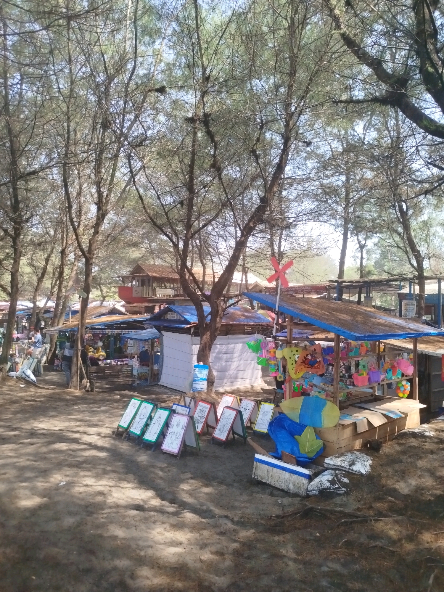 The image depicts a vibrant marketplace set up beneath a canopy of trees, likely on a beach due to the sandy ground. The scene is located in Kenoyajaya, Indonesia, and captures a lively atmosphere with various stalls selling colorful items.
In the foreground, several small, easel-like displays are set up, each with a drawing or painting on it. These appear to be for sale, perhaps as art or souvenirs. Behind these displays, a stall is laden with a variety of merchandise. Brightly colored toys, buckets, and possibly kites are visible, suggesting a focus on children's items or beach accessories. A small red windmill with four blades is mounted on a pole above one of the stalls, adding a playful touch to the scene.
Further back, more stalls and structures can be seen, with blue and brown awnings providing shade. In the background, a two-story building with a red base and wooden upper level stands out, possibly a cafe or restaurant. The trees overhead are mostly bare, with some sparse green foliage, indicating a dry season or the type of trees common to the area. The ground is covered in sand and some scattered debris.
Several individuals are present, though most are not clearly visible or are engaged in activities that are difficult to discern from this distance. One person in dark clothing appears to be working at a stall. The overall impression is of a bustling outdoor market catering to visitors in a recreational area.