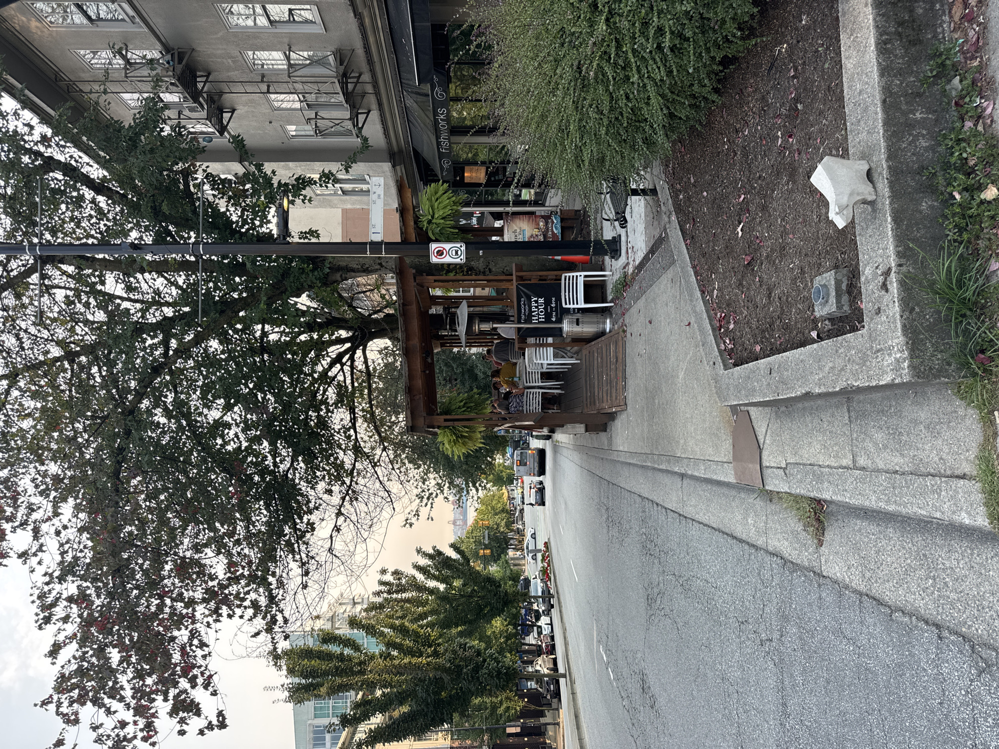 An outdoor seating area for a restaurant, likely in North Vancouver, Canada, is depicted on the right side of the image. A covered wooden structure with tables and chairs is set up on a raised platform next to a street. Several people are seated at the tables, some appearing to be engaged in conversation. A "Happy Hour" sign is visible, indicating the restaurant's offerings. The street in front of the restaurant is paved and has visible lane markings. Cars are parked along the sidewalk and driving on the street. To the left of the street, a building with multiple windows and fire escapes is visible. Trees with lush green foliage line the street and the outdoor seating area, suggesting it is a temperate season. A street lamp and a traffic signal are also present. The overall scene captures a typical urban streetscape with outdoor dining.