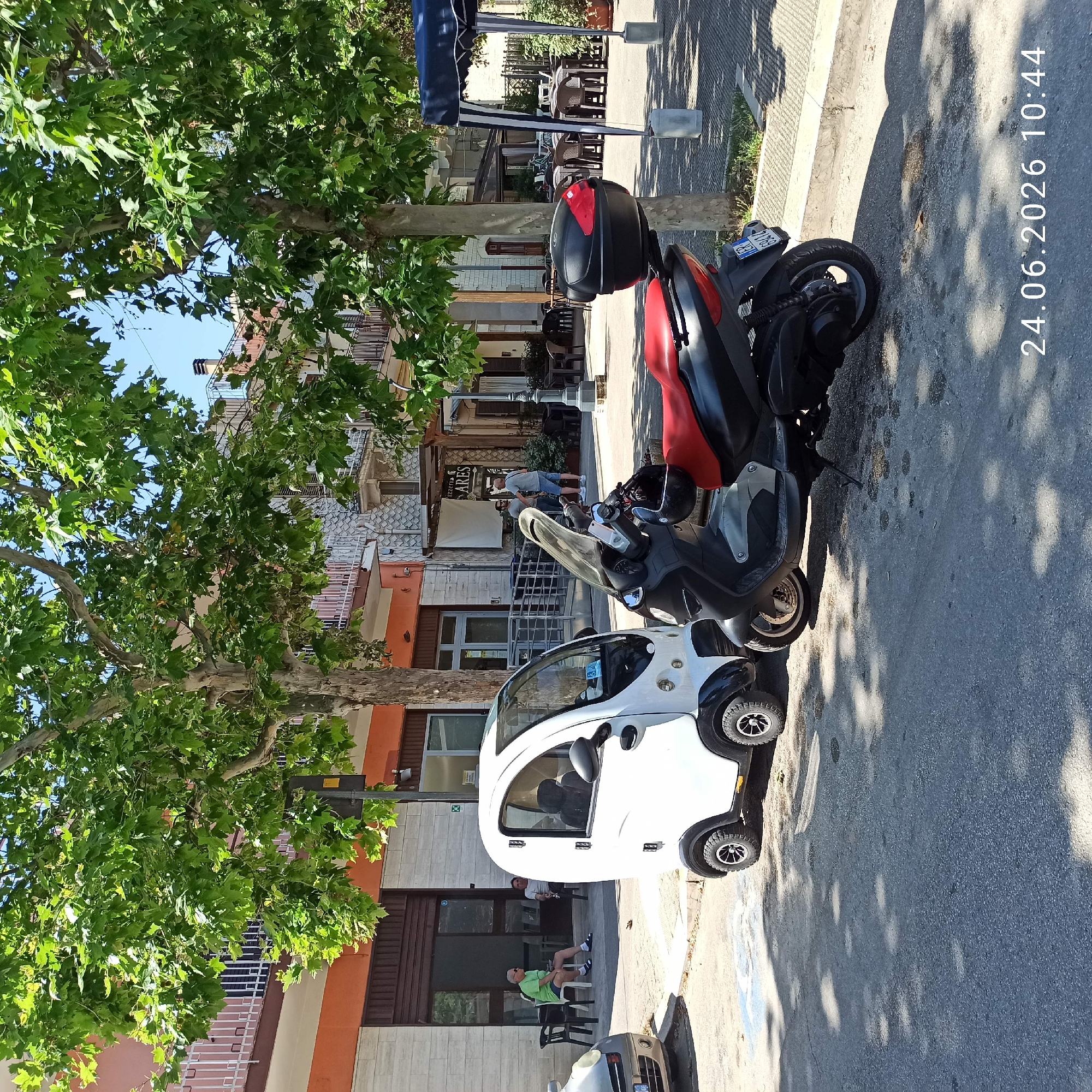 The image shows a street scene in Bitonto, Italy, on a sunny day, likely in the late morning judging by the shadows. In the foreground, a white, three-wheeled vehicle, similar to a small car or enclosed scooter, is parked on the asphalt. Behind it, a red and black scooter is also parked. Both vehicles are on the side of a street lined with buildings.
On the sidewalk in front of a building, two men are seated on chairs. One man, wearing a green t-shirt and shorts, is relaxed, leaning back. The other man, wearing a grey t-shirt and shorts, is also seated. The buildings have a mix of architectural styles, with some featuring balconies and orange and cream colored facades. Trees with lush green leaves provide shade, casting dappled shadows on the street and sidewalk.
Further down the street, outdoor seating for a cafe or restaurant is visible, with several tables and chairs set up under a blue awning. The overall atmosphere appears to be calm and relaxed, typical of a pleasant day in a European town.
Visible text includes a license plate on the scooter that reads "FR 872174" and the date "24.06.2026 10:44" is superimposed on the image. There is also some text on a sign above the cafe seating area, which appears to read "PIZZERIA" and "ARIAS".