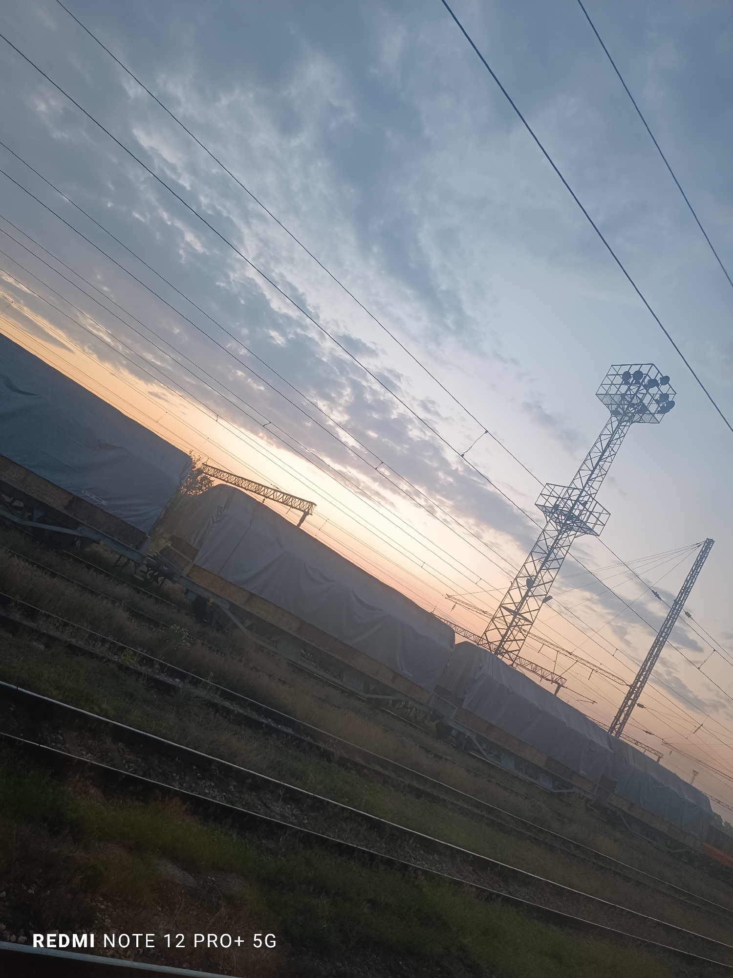 This image captures a tranquil railway scene during either sunrise or sunset, set against a sky painted with warm orange and yellow hues near the horizon, gradually fading into cool blues and grays further above. Scattered clouds are present, adding texture to the sky.
A prominent feature is a long line of stationary freight train cars, each covered with a light-colored, possibly gray or white, tarp or canvas. These cars rest on multiple parallel railway tracks that stretch across the lower half of the frame and into the distance. Grass and sparse vegetation grow between the tracks.
Overhead, numerous thin power lines, indicative of an electrified railway system, crisscross the sky. To the right, a tall metal lattice tower stands out, featuring a large, multi-faceted structure at its top, likely an array of floodlights or communication equipment. Another, thinner lattice support structure is partially visible further to the right.
The scene depicts a railway yard or siding, characteristic of a significant transportation hub such as Kaspichan, Bulgaria. No people or active events are visible, contributing to a sense of stillness. The photograph is taken at an oblique angle, creating a diagonal composition. A watermark "REDMI NOTE 12 PRO+ 5G" is visible in the bottom-left corner.