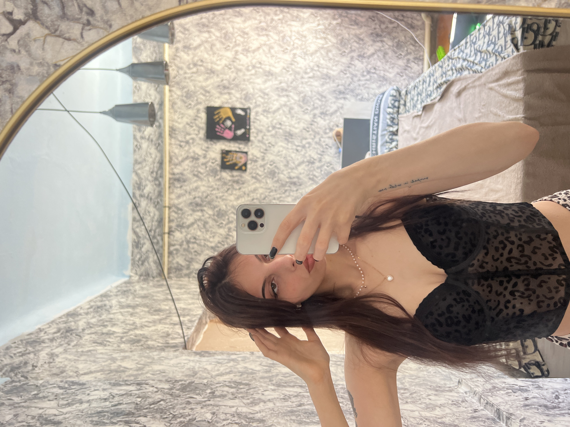 A young woman with long, dark hair and dark nail polish is taking a selfie in a mirror. She is wearing a black leopard-print corset top and leopard-print bottoms. She is also wearing a pearl necklace with a heart-shaped pendant. Her right arm is raised, and her hand is touching her hair. There is a tattoo on her left arm. In the mirror's reflection, we can see a bed with patterned bedding, several hanging light fixtures, and two framed handprints on a textured wallpapered wall. The room appears to be a bedroom.