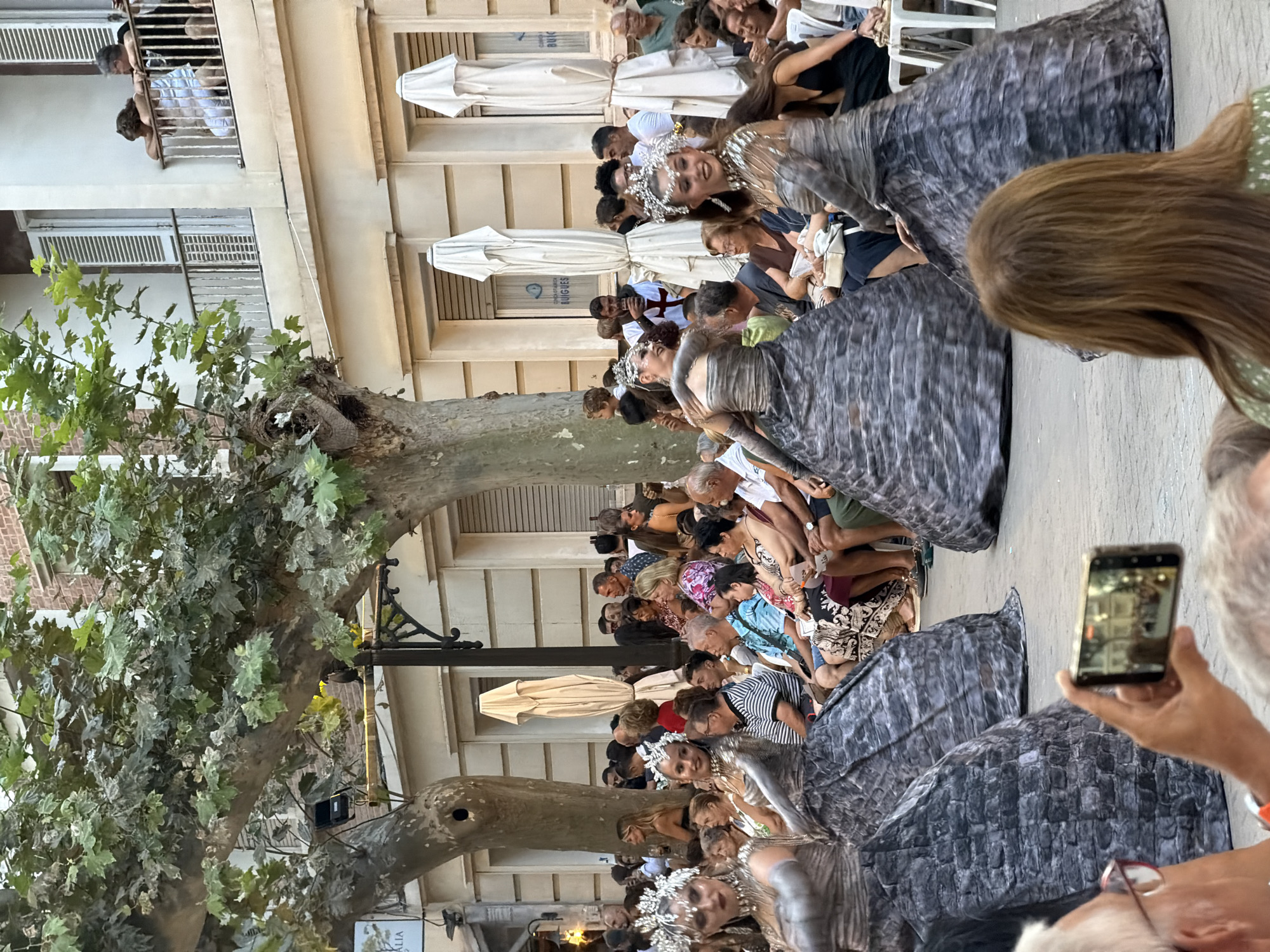 The image captures an outdoor event, likely a performance or parade, taking place on a street in the city of Dénia, Spain. In the foreground, two performers are dressed in elaborate, gray, textured dresses that resemble stone or brickwork, adorned with silver headpieces. They are positioned in a way that suggests they are either moving or posing for the audience.
The audience is seated and standing around the performers, with many people looking towards the performance. Some individuals in the audience are holding up mobile phones, presumably to capture photos or videos of the event. The background features buildings with balconies and windows, and large trees with lush green foliage line the street, casting shadows on the scene. The presence of white umbrellas suggests that this event might be taking place during the day, possibly in a public square or street that has been temporarily set up for the occasion. The architecture and the style of the buildings, combined with the trees, suggest a European setting, consistent with Dénia, Spain.