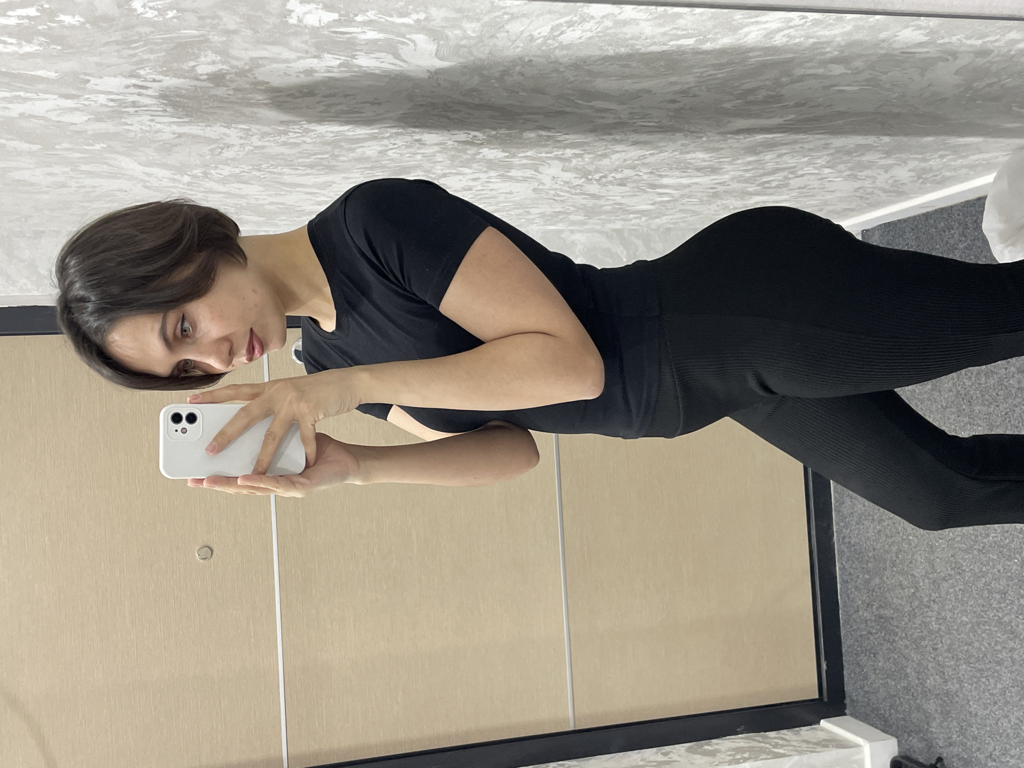 A woman with short brown hair wearing black athletic clothing is taking a selfie in front of a full-length mirror. She is holding a white smartphone. The environment appears to be a room with light beige walls and gray speckled flooring. There is no indication of the city of Izhevsk, Russia.