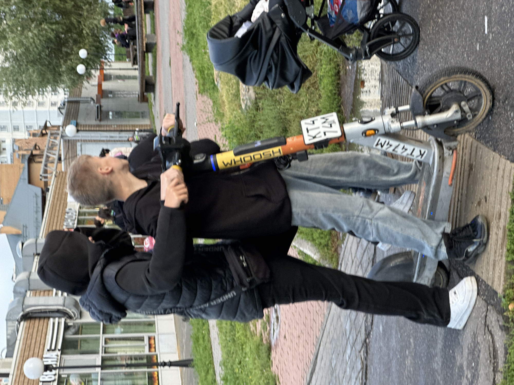 Two individuals are on an electric scooter on a paved walkway in Podolsk, Russia, near buildings with commercial storefronts. The person in the foreground, on the right, is a youth with short blonde hair, wearing a black hoodie and light blue jeans. They are operating the scooter. Behind them, to the left, is another person wearing a black hooded jacket and black pants, with a black crossbody bag over their shoulder. This person's face is obscured by their hood. The scooter is orange and silver, with the "WHOOSH" logo and license plate "KT4 74M" visible. To the right of the scooter is a black stroller with a covered baby seat. The environment shows green grass, a brick-patterned walkway, and buildings in the background, including a multi-story residential building. The sky is overcast.