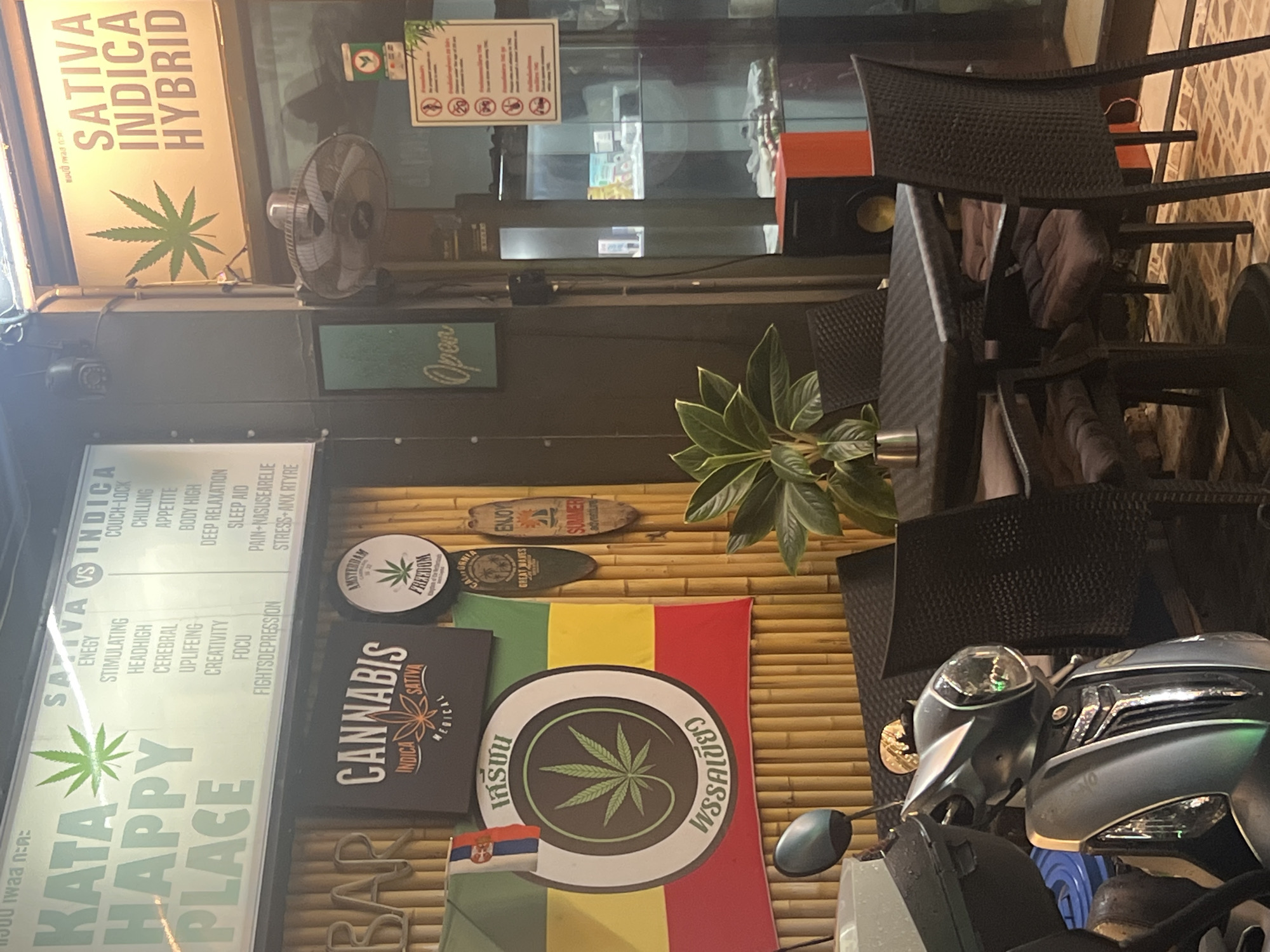The image depicts the exterior of a cannabis shop in Karon, Thailand. The scene is dominated by various signs and decorations related to cannabis. A large sign on the left reads "KATA HAPPY PLACE" with "SATIVA VS INDICA" above it, listing effects and characteristics of different cannabis strains. Another sign displays "SATIVA INDICA HYBRID".
Various circular and rectangular signs are displayed, including one that says "AMSTERDAM FREEDOM" and another with "CANANIS SATIVA INDICA MEDICAL". A flag with red, yellow, and green stripes, reminiscent of Rastafarian colors, is prominently displayed with a circular cannabis leaf logo. Smaller flags, including one from Serbia, are also visible.
The setting appears to be an outdoor storefront with bamboo-like paneling in the background. Furniture, including chairs, is visible, and a fan is mounted on the wall. A plant with large green leaves adds a touch of nature to the scene. In the foreground, parts of a scooter are visible, suggesting this is a street-level establishment.
The lighting suggests it might be daytime or early evening, with some artificial light sources contributing to the illumination. The overall impression is that of a casual, possibly tourist-oriented establishment catering to cannabis enthusiasts, with a strong visual emphasis on cannabis culture and information. The text elements are a key feature, providing information about cannabis strains and the shop itself.