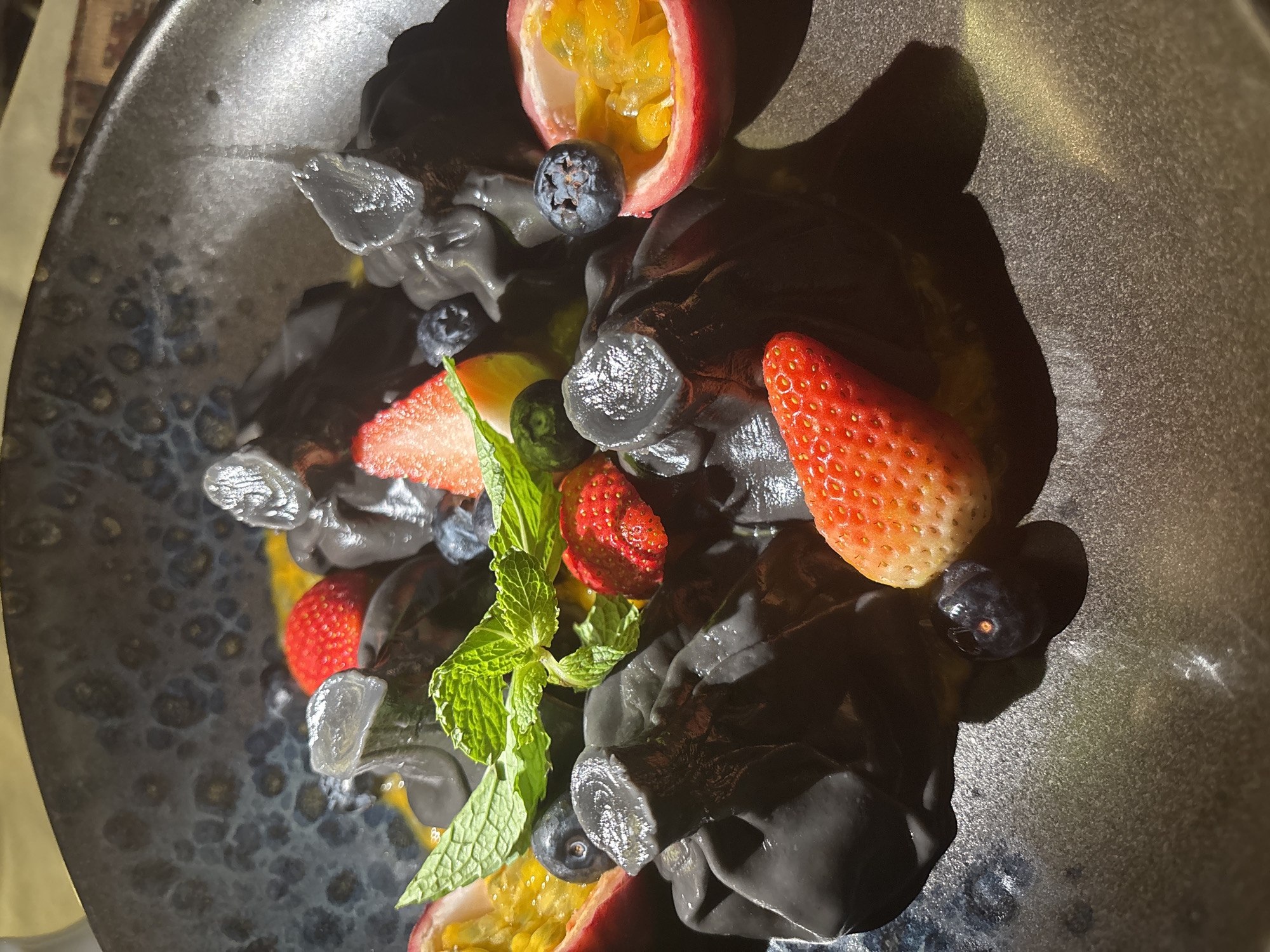 A close-up, high-angle shot shows a dessert dish, potentially photographed indoors, in Choeng Thale, Thailand. The dish contains black, oddly shaped pasta. The pasta is garnished with halved passion fruit, blueberries, sliced strawberries, and a sprig of mint. The plate itself is dark gray with a textured, possibly ceramic or stoneware, surface. The lighting creates strong shadows, emphasizing the textures and shapes of the food. No people are visible in the frame.