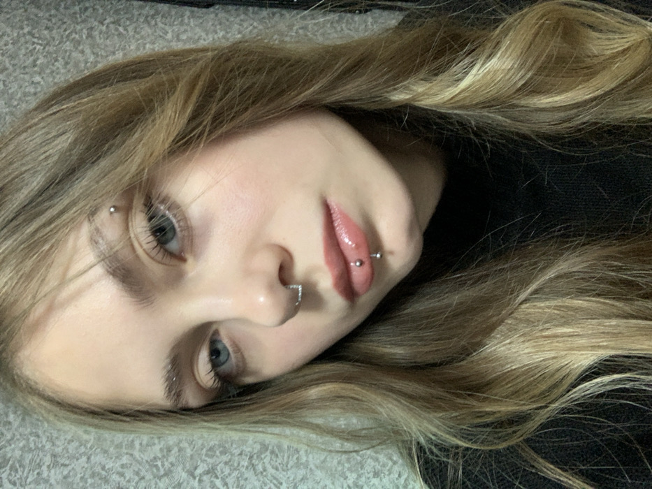 A fair-skinned young woman with blonde-highlighted brown hair is captured in a close-up, slightly tilted view. Her features are accentuated by several piercings: one through the eyebrow, a ring in her nose, and a bar through her lower lip. She gazes towards the camera with light-colored eyes. Her lips are glossy, and she appears to be lying down, with part of her dark-colored garment visible. The image is softly lit.