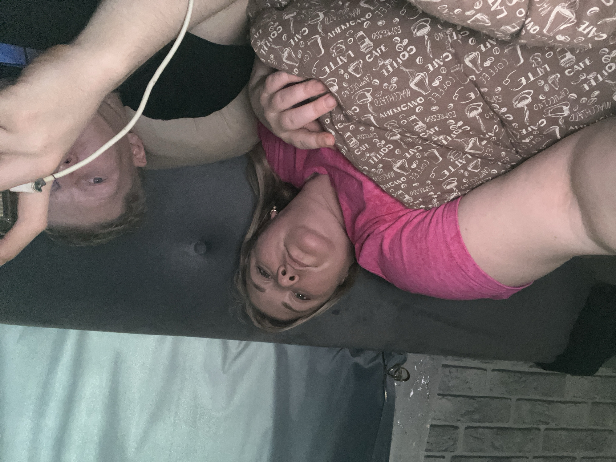 Two people are lying on a gray fabric surface, possibly a couch or bed. A woman with blonde hair and a pink shirt is positioned on the right side of the frame, her head facing the camera. To her left, a brown pillow with a coffee-themed print is visible. On the left side of the frame, a man with fair skin and short hair is present, partially obscured by a white cable. He appears to be looking at a device in his hand. The backdrop consists of a light blue curtain and a brick-patterned wall. There is no information to identify that the photo was taken in Vesyoloye, Russia.