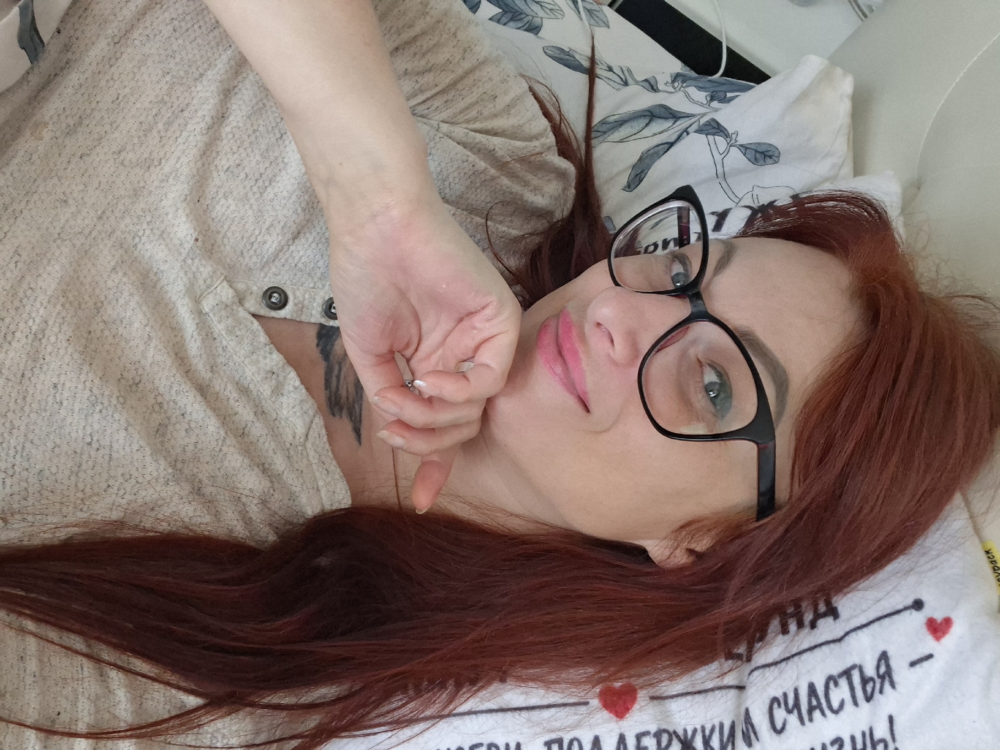 A woman with long, auburn hair and black-rimmed glasses lies on her side, propping her head up with her hand. She is wearing a light-colored, textured shirt with two visible buttons. Her lips are painted a bright pink. A tattoo is visible on her left shoulder blade. She is positioned on a bed with a white pillow featuring a floral pattern and text in Cyrillic. The background is indistinct but suggests an indoor setting. The image appears to be a selfie taken from a close-up, high-angle perspective. There is no information about the city of Myadzyel, Belarus present in the image itself.