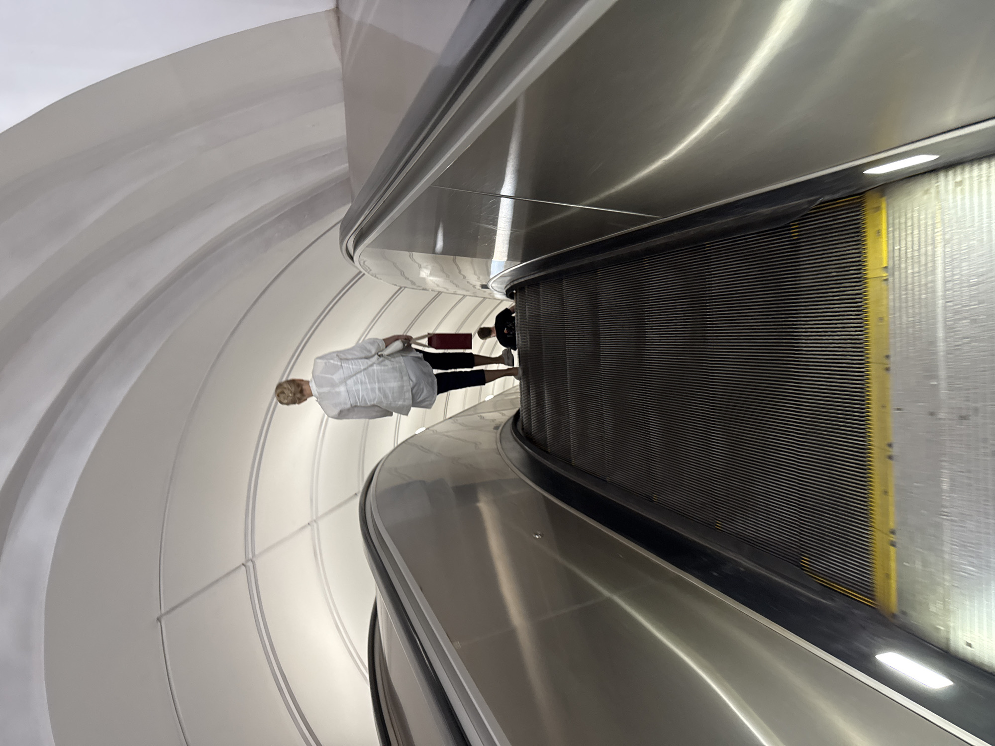 The image shows a wide escalator with metal sides and black treads, illuminated by embedded lights. A woman with blonde hair pulled back is walking up the escalator, wearing a white shirt and dark pants, carrying a red bag. Further up the escalator, a person is seated. The escalator is set within a large, arching tunnel, which is likely in the Moscow Metro system, known for its grand architectural designs. The overall impression is one of movement and public transit within an impressive subterranean environment.