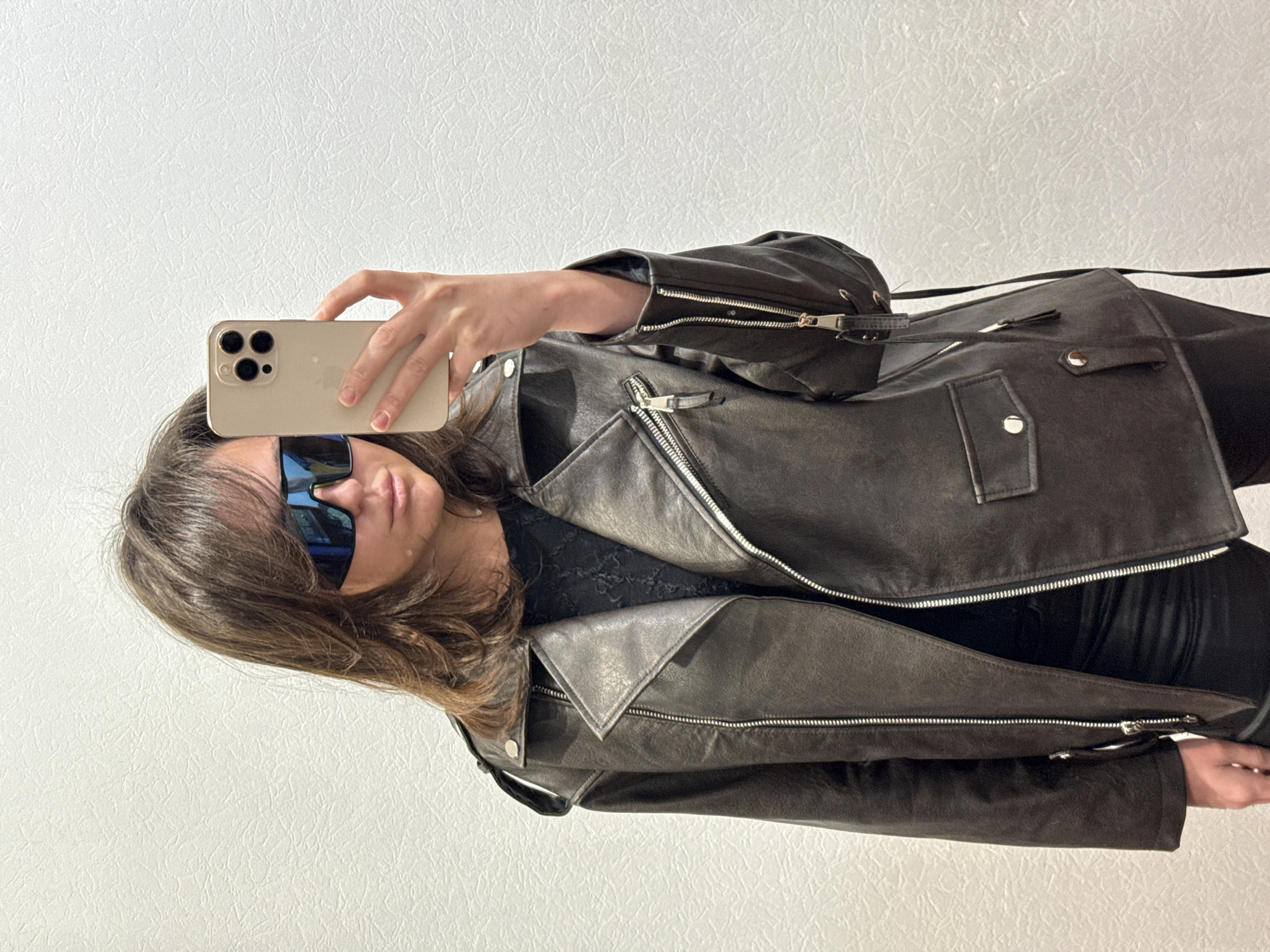 A woman in a black leather jacket and sunglasses takes a selfie with a gold iPhone. The background is a white wall. The image is oriented sideways. The location is not discernible.