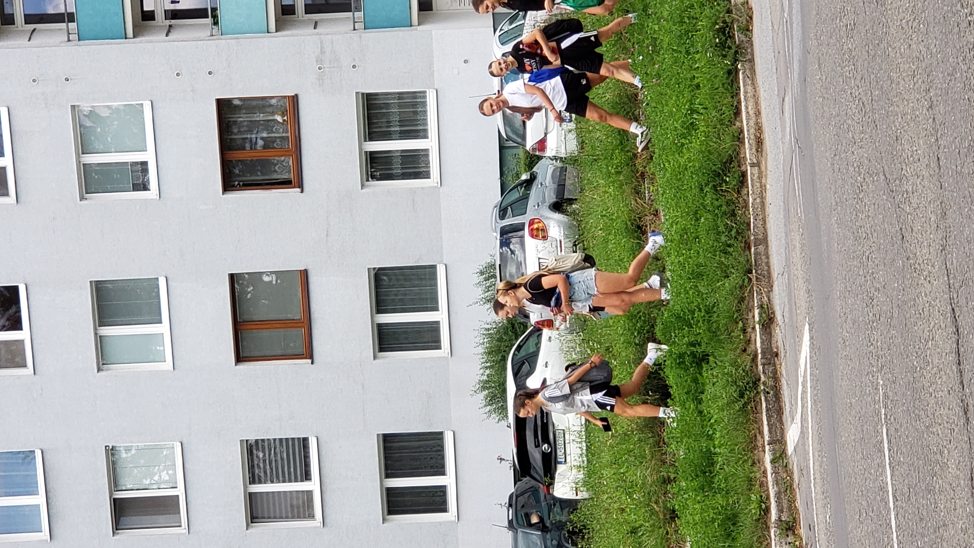 In Prešov, Slovakia, a group of five young women walks along a grassy area adjacent to a road. The women, appearing to be teenagers or young adults, are dressed in athletic attire, including shorts and t-shirts. They are walking from left to right in the frame. The background features a multi-story apartment building with numerous windows, and parked cars are visible behind the women. The environment suggests an urban residential area.