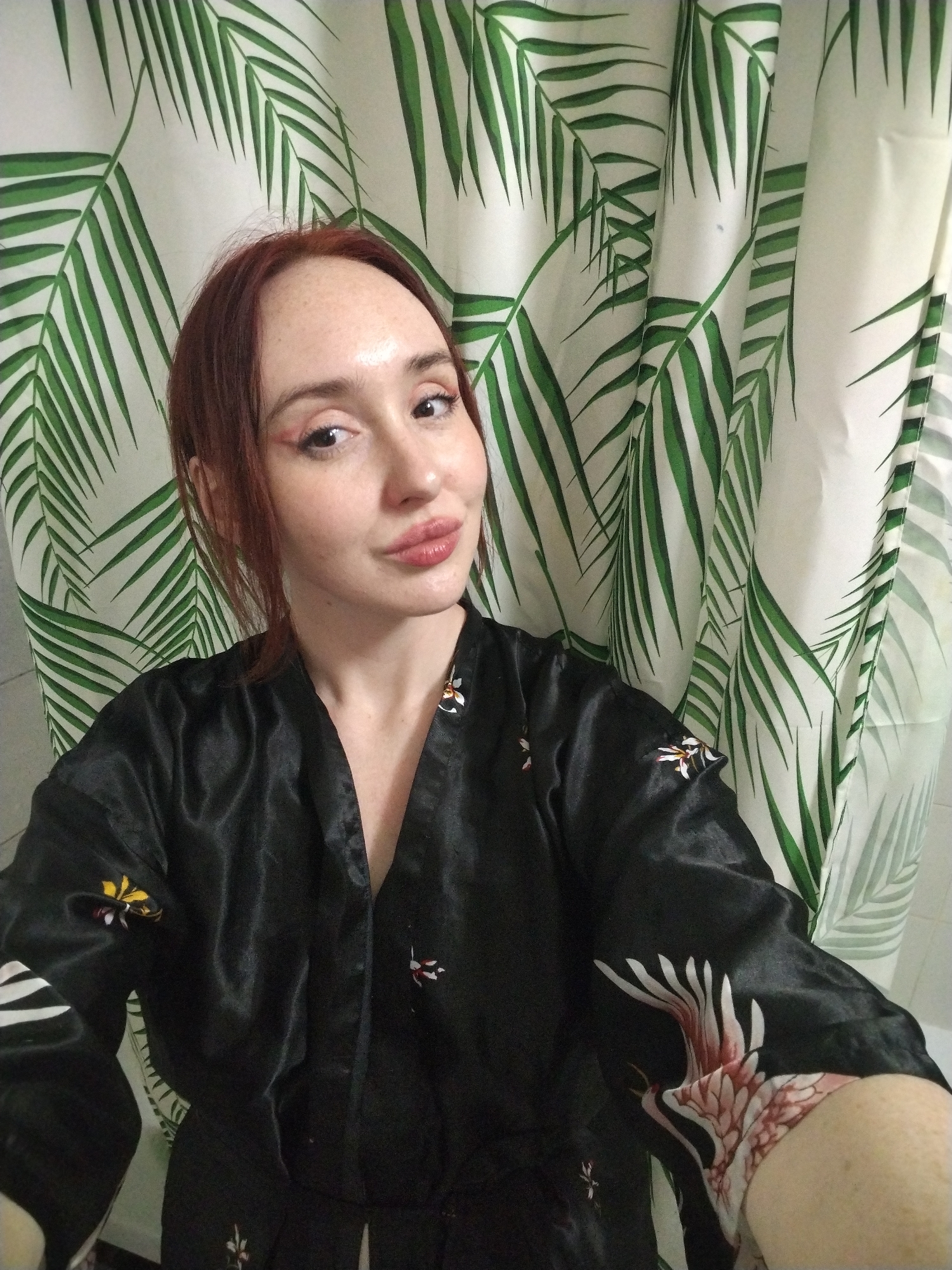 A selfie of a fair-skinned woman with red hair, rosy makeup, and freckles wearing a black silk robe with floral embroidery. The background is a white shower curtain with green palm leaf patterns. The location is not discernible from the image and cannot be confirmed as Izhevsk, Russia.