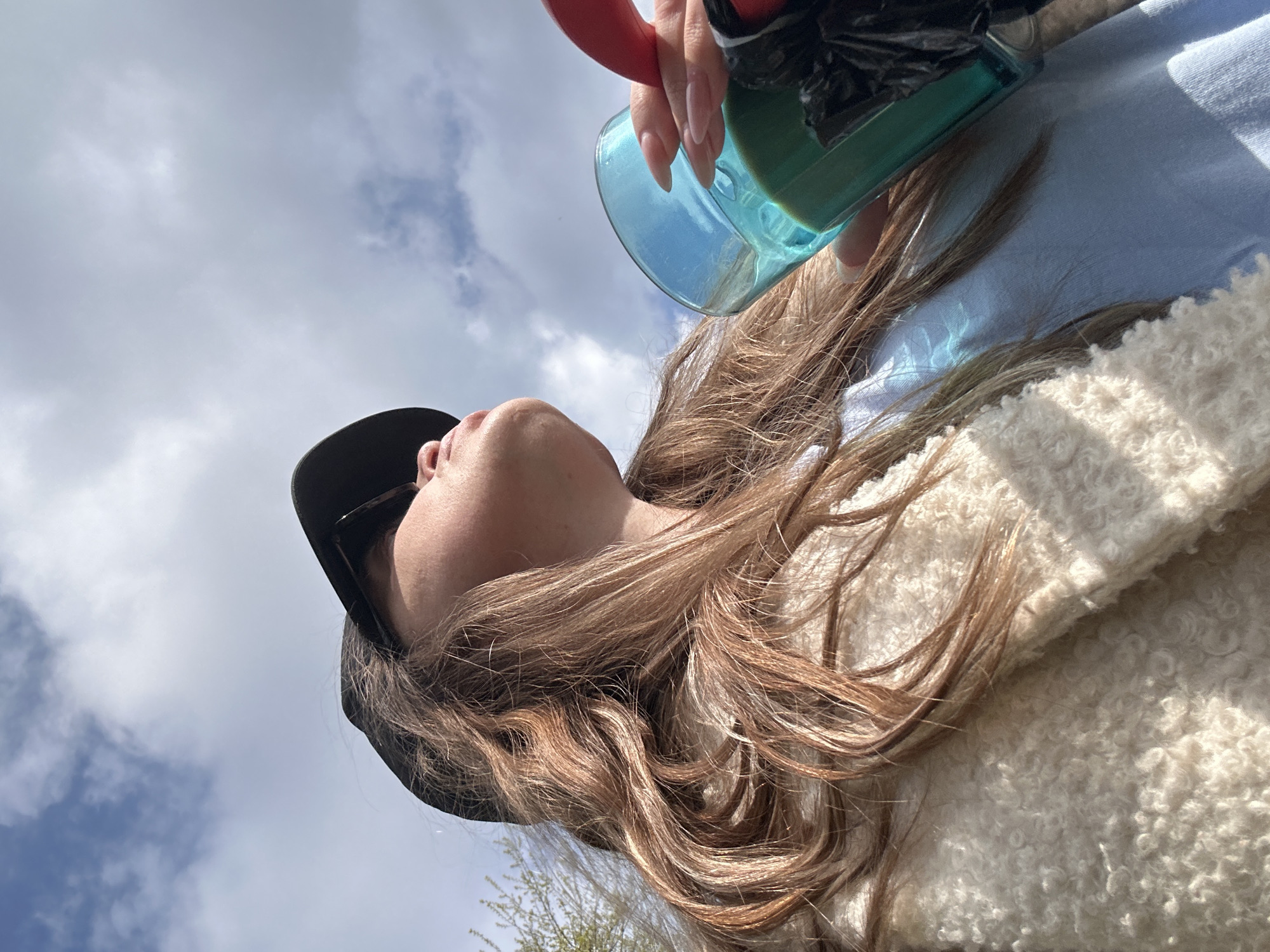 A woman wearing a black cap and a fuzzy, light-colored coat is looking up at the sky, holding a blue glass filled with a dark liquid. The sky is visible and partly cloudy. This photo was likely taken from a low angle. The woman appears to be on a sunny day, and the location of Tbilisi, Georgia is not discernible from the image.