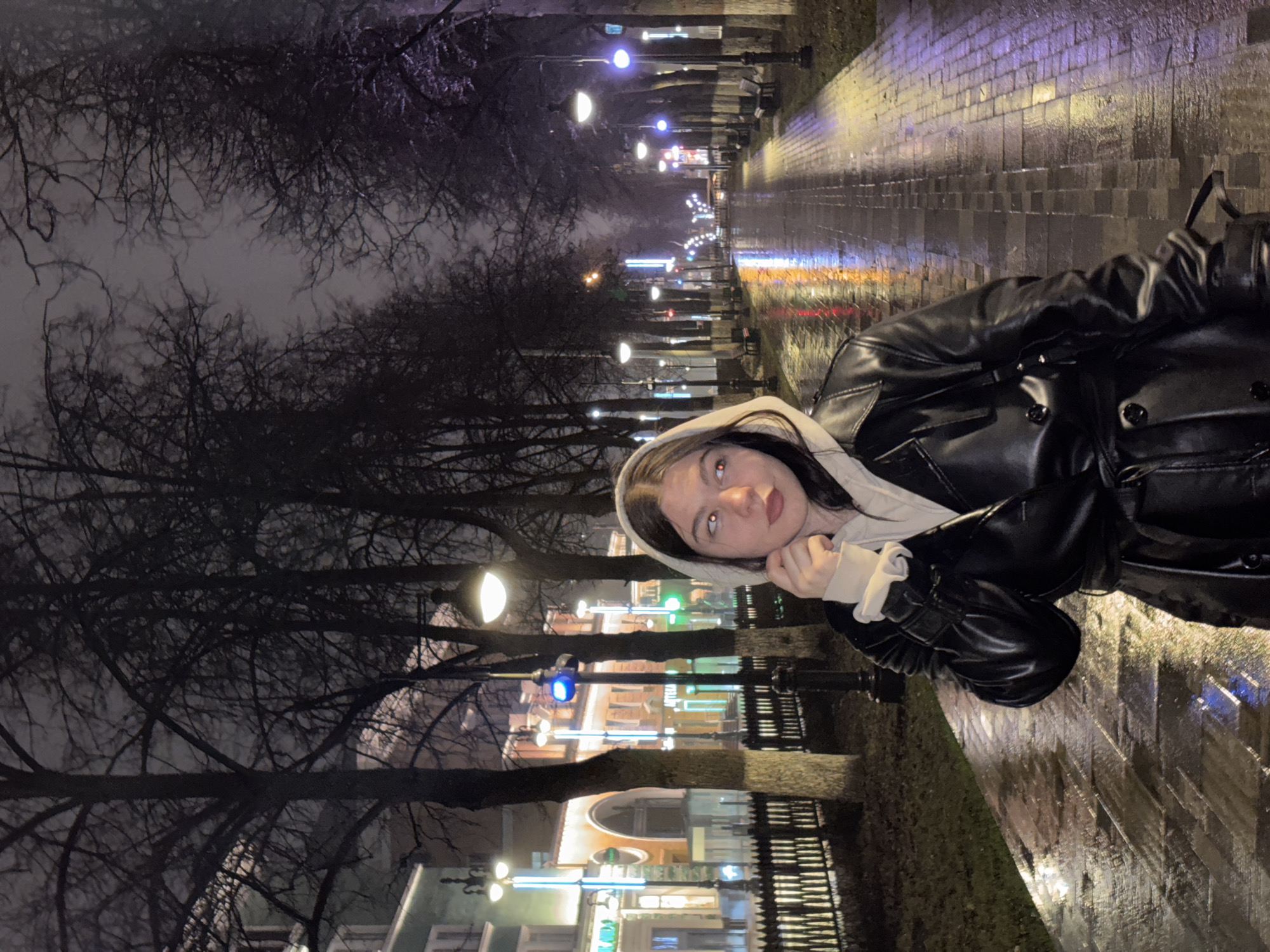 A young woman with pale skin, dark hair, and dark eyes is depicted in the image. She wears a light-colored hoodie under a black leather jacket and rests her chin on her hand, looking upward. She is standing on a wet, paved walkway.
The location is a public thoroughfare in Perm, Russia, at night, likely after rainfall. Bare trees line the walkway on the left, and buildings with illuminated facades can be seen behind them. Light posts with glowing lamps are spaced along the walkway, their reflections visible on the wet pavement, which is likely cobblestone. In the distance, more lights illuminate the street. The overall impression is of a quiet, damp evening in an urban setting.