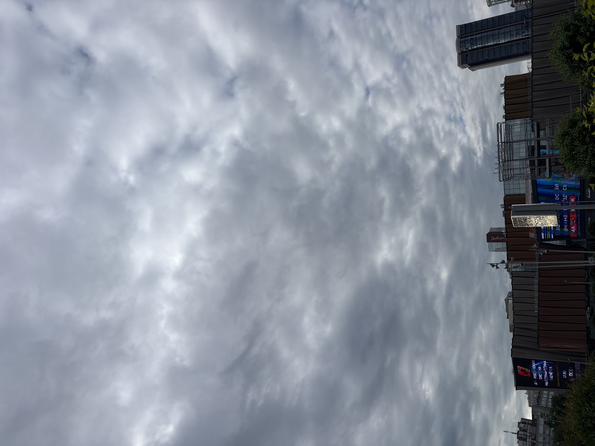 The image depicts an urban landscape under a heavily overcast sky in Perm, Russia. The upper two-thirds of the frame are dominated by a dense, grey cloud cover, indicating an overcast day with no direct sunlight visible.
The lower third of the image shows a complex of modern buildings. On the far left, a light-colored multi-story building is partially visible behind some trees. In the mid-ground, a large commercial building complex features a textured, possibly brown or dark-toned facade with vertical paneling. A prominent, large outdoor LED screen billboard on this complex displays Cyrillic text, reading "A7 - РОССИЙСКАЯ СИСТЕМА МЕЖДУНАРОДНЫХ РАСЧЕТОВ" (A7 - RUSSIAN SYSTEM OF INTERNATIONAL SETTLEMENTS), along with "A7.RU" and "ПСБ". Further to the right on the same complex, another large LED screen is partially visible, displaying vibrant colors and more Cyrillic text. A section of scaffolding or exposed structure is visible on a lower portion of this central building.
To the right of the central complex, a tall, slender skyscraper with dark glass windows rises prominently. Above the central building, a red "Radisson" sign is visible on a rooftop structure, confirming the location as near the Radisson Hotel Perm. A vertical pole equipped with multiple loudspeakers or light fixtures stands between sections of the buildings. Dark green foliage from trees or bushes is visible along the very bottom edge of the image, partially obscuring the base of the structures. No people or discernible activities are visible within the frame.
