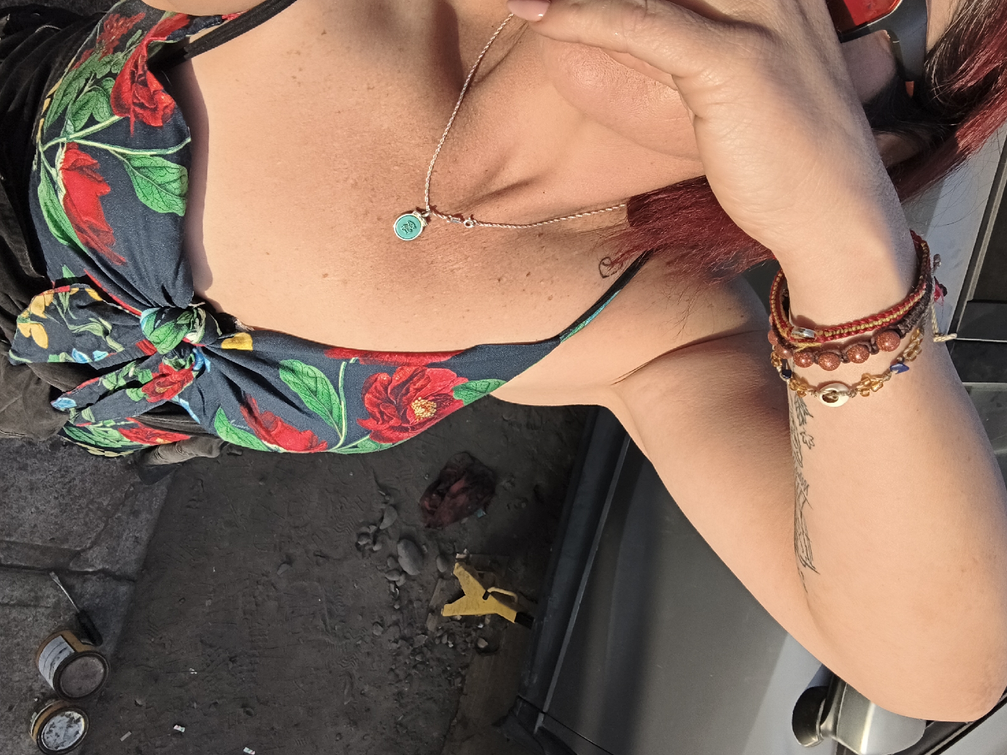 An overhead shot presents a view of a person, likely female, with red hair and sunglasses perched on their head. The person is wearing a floral patterned top and multiple bracelets on their left wrist. A silver necklace with a turquoise pendant adorns their neck. The setting appears to be outdoors. On the ground, there are various items, including what looks like a yellow clamp, a dark-colored cloth or bag, and two small paint cans. The ground surface appears to be concrete or stone. The image does not provide enough information to place it in the city of Arica, Chile, with certainty.