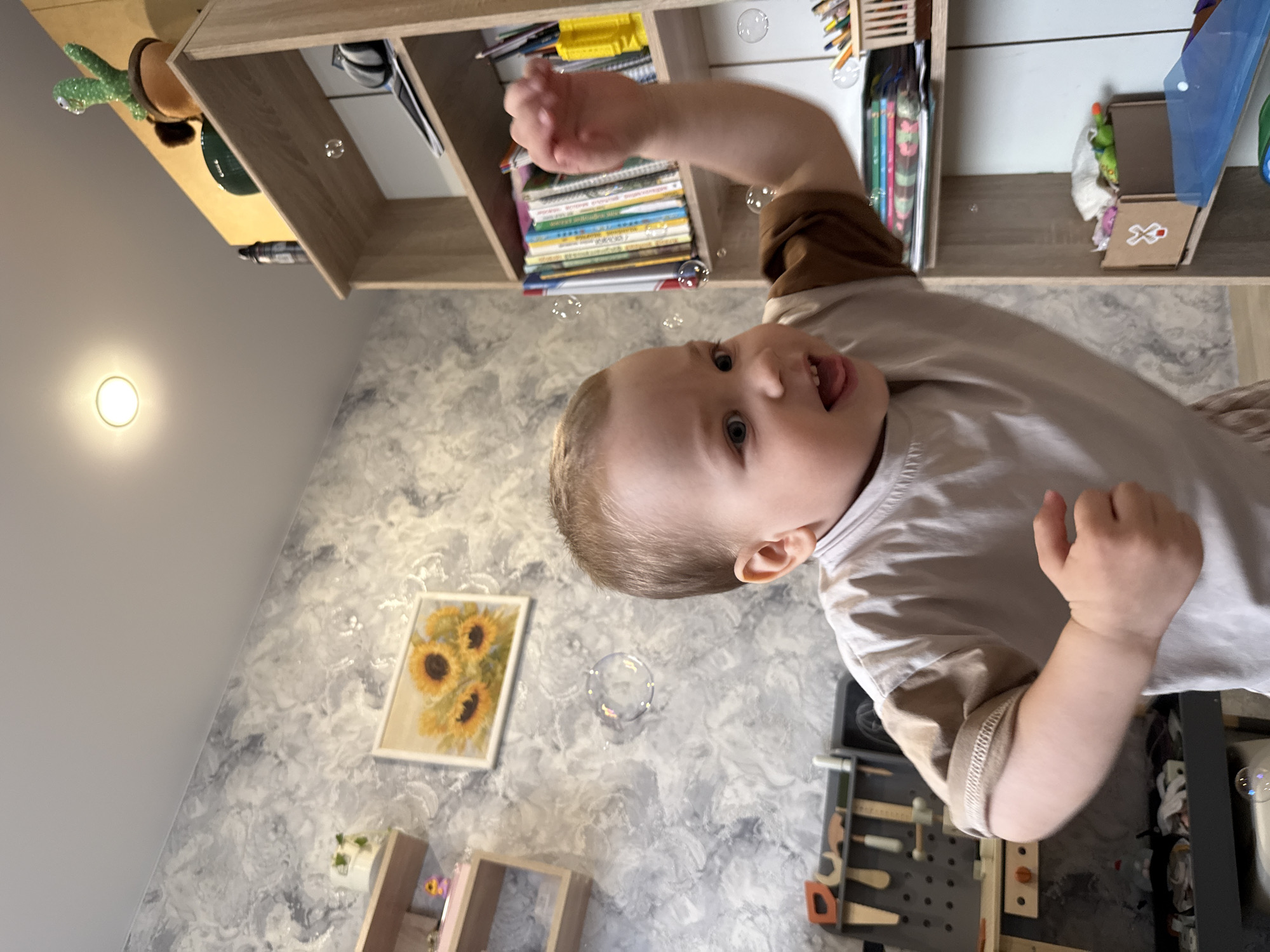 A young child is photographed indoors, likely in a child's room. The child is facing the camera with their mouth open and one arm raised. Bubbles are floating around them. In the background, there is a shelf containing books, art supplies, and a cactus. On the wall, there is a painting of sunflowers. Below the painting, there is a small wooden shelf. The wall has a marbled texture. There is a light on the wall. There is a tool set and other toys next to the child. The exact location within Rusanovo, Russia, cannot be determined from the image.