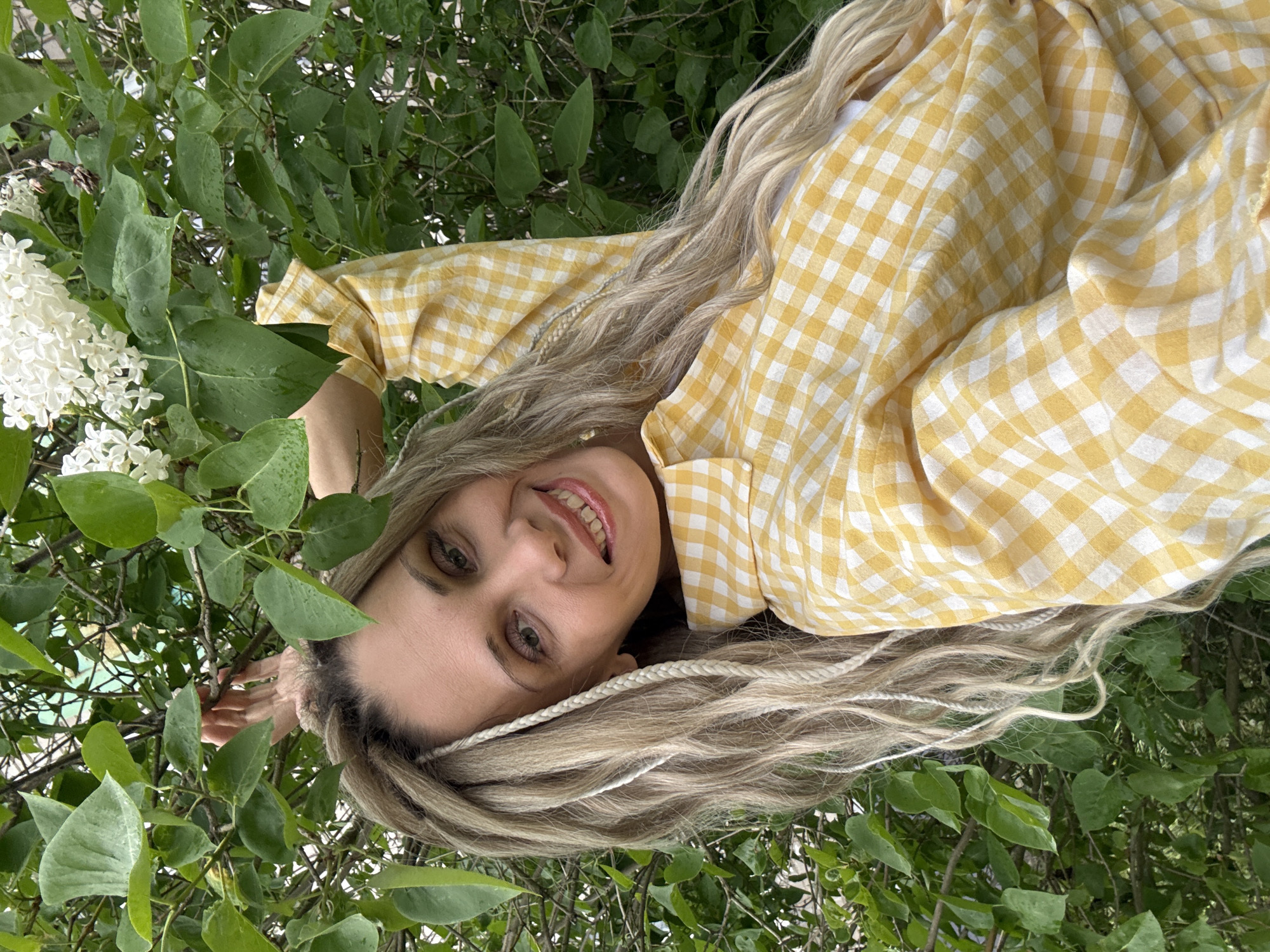A woman with long, light-blonde hair styled with thin, white braids lies among green leaves and white flowers, likely lilac bushes. She is wearing a yellow and white gingham shirt. The photo appears to be taken from an upward angle, with the woman looking down at the camera, smiling. This image appears to have been taken outside in the city of Rusanovo, Russia.