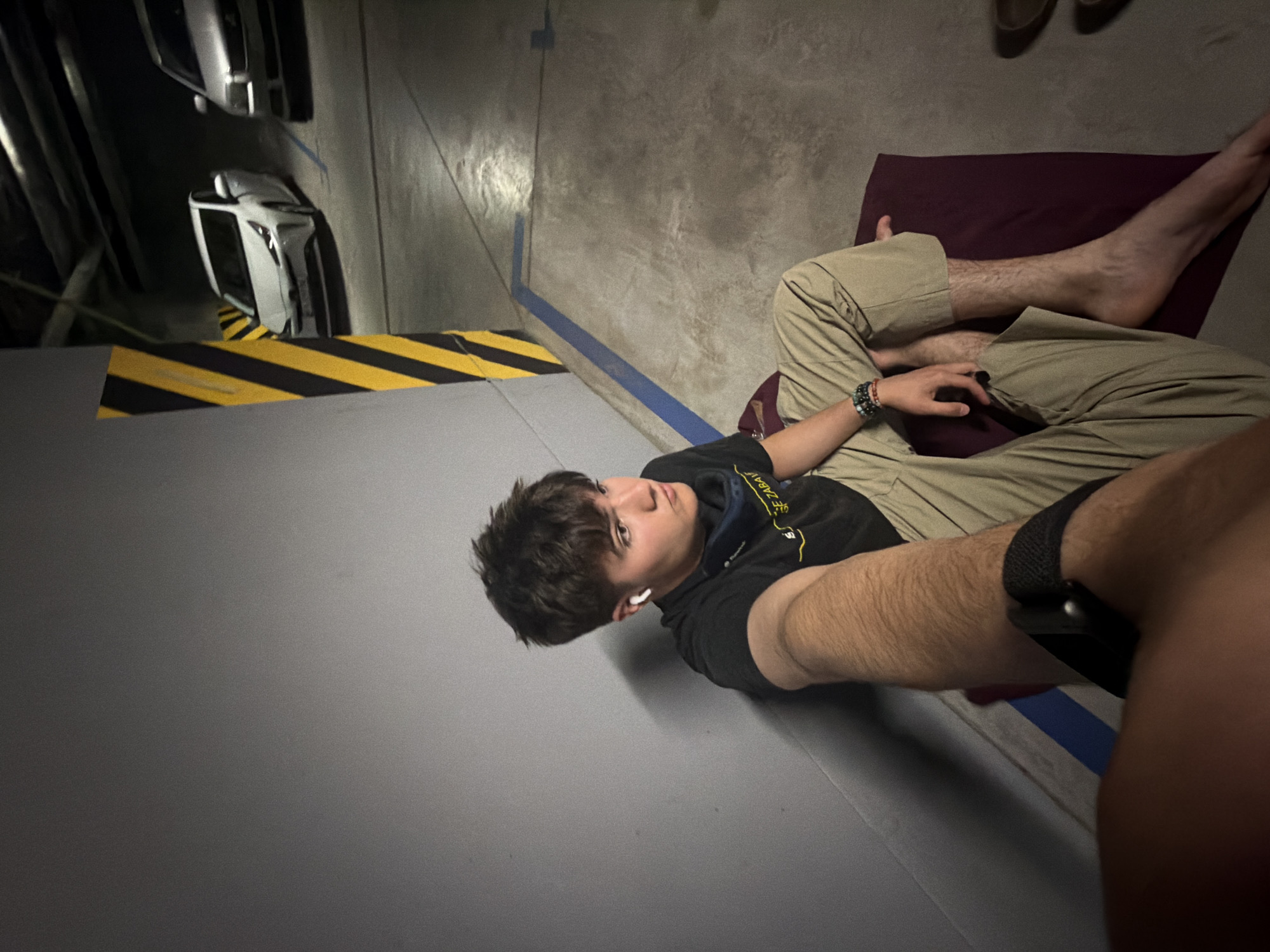 A young male with brown hair, wearing a black t-shirt, khaki pants, and white earbuds, sits on a maroon mat in a dimly lit parking garage. He is leaning against a grey concrete wall, with a yellow and black striped pillar to his left. In the background, two cars are parked, a white sedan and a silver sedan. A blue line is painted on the floor. His left arm is extended towards the camera, and he is wearing a watch on his left wrist and bracelets on his right wrist. The scene appears to be a candid shot taken in a public space. The location is identified as a parking garage in Kyiv, Ukraine.