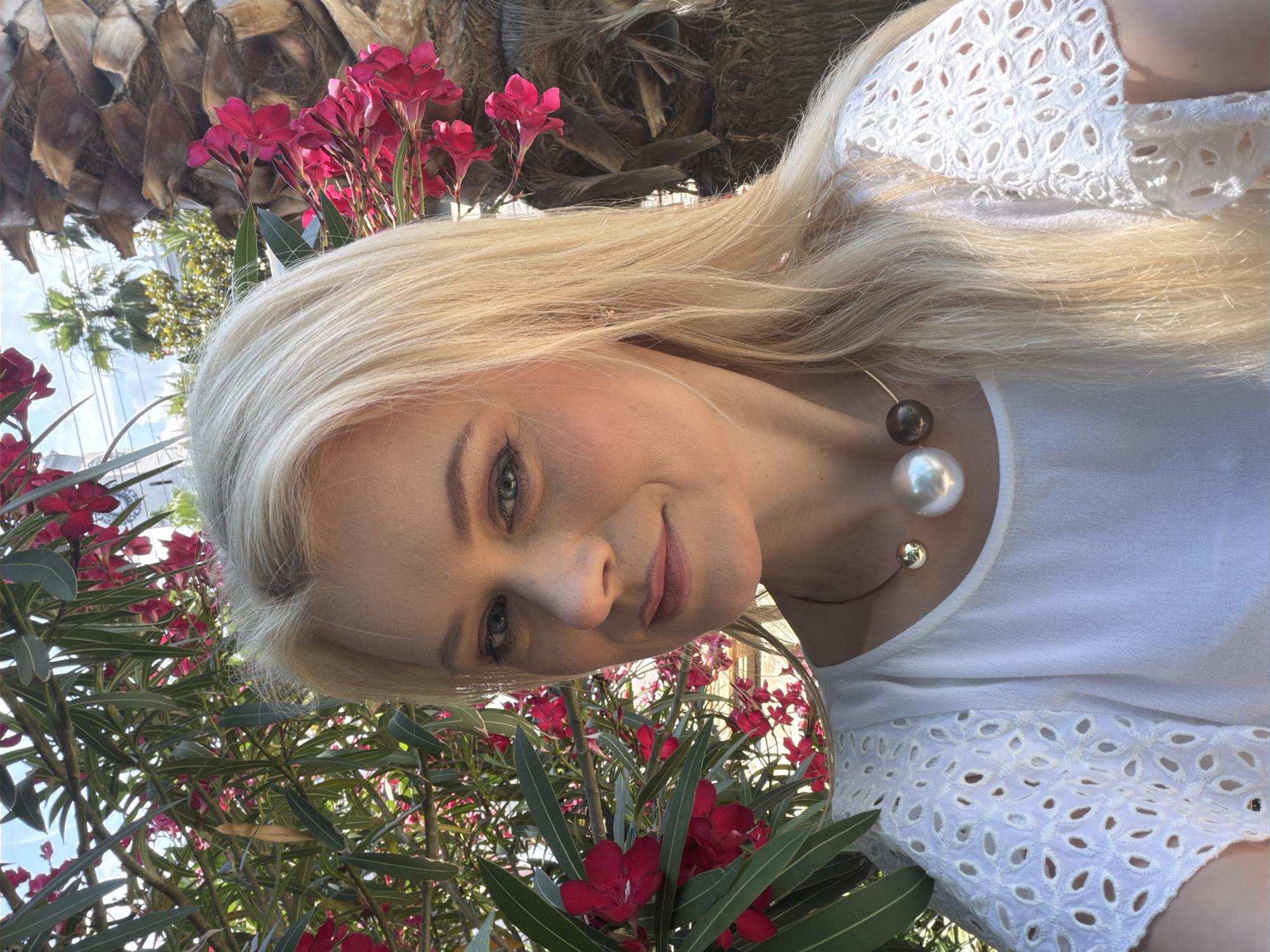 A blonde woman with blue eyes and fair skin is pictured outdoors, posing for a selfie. She is wearing a white blouse with eyelet detailing on the sleeves and a statement necklace featuring a large pearl and two dark spheres. The background features lush green foliage with pink flowers and a palm tree, suggesting a warm, possibly tropical or subtropical climate. The lighting indicates it is daytime. The specific location in the city of Sochi, Russia, is not discernible from the visual elements alone, as such flora and palm trees are common in the coastal regions of this city.