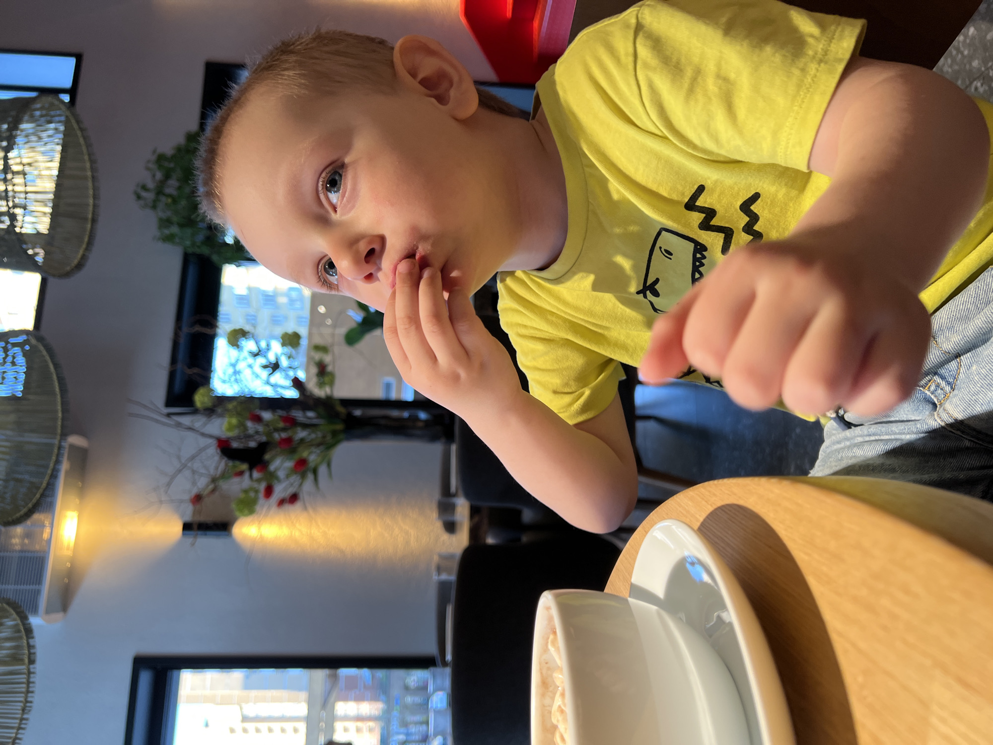 A young, fair-skinned boy in a yellow t-shirt with a cartoon dinosaur graphic is sitting at a round, light-colored wooden table. He has blond hair and blue eyes, and is holding his fingers to his mouth as if eating something. A white cup and saucer are on the table in front of him. The scene appears to be indoors, possibly a cafe or restaurant, with a window in the background showing a cityscape, and floral arrangement on the table. Considering the reference, it is likely this scene is taking place in the city of Mytishchi, Russia. Woven light fixtures are visible hanging from the ceiling. The photograph is taken at a slightly tilted angle.