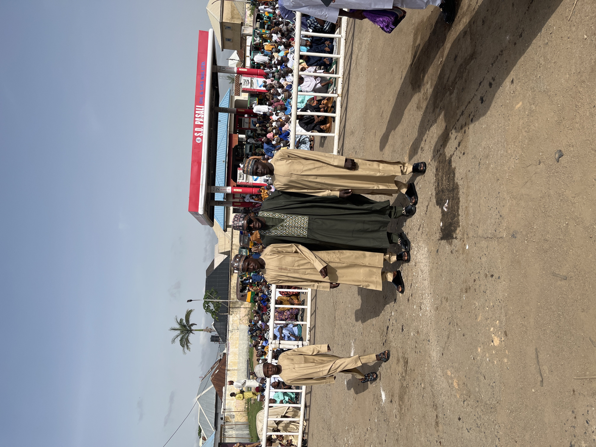 The image shows four men in traditional Islamic attire standing in a row on a paved surface near a gas station identified as "S.D. PASALI" in Jos, Nigeria. A crowd of people is seated behind a white fence in the background. A palm tree is visible to the left, and a clear blue sky is above.